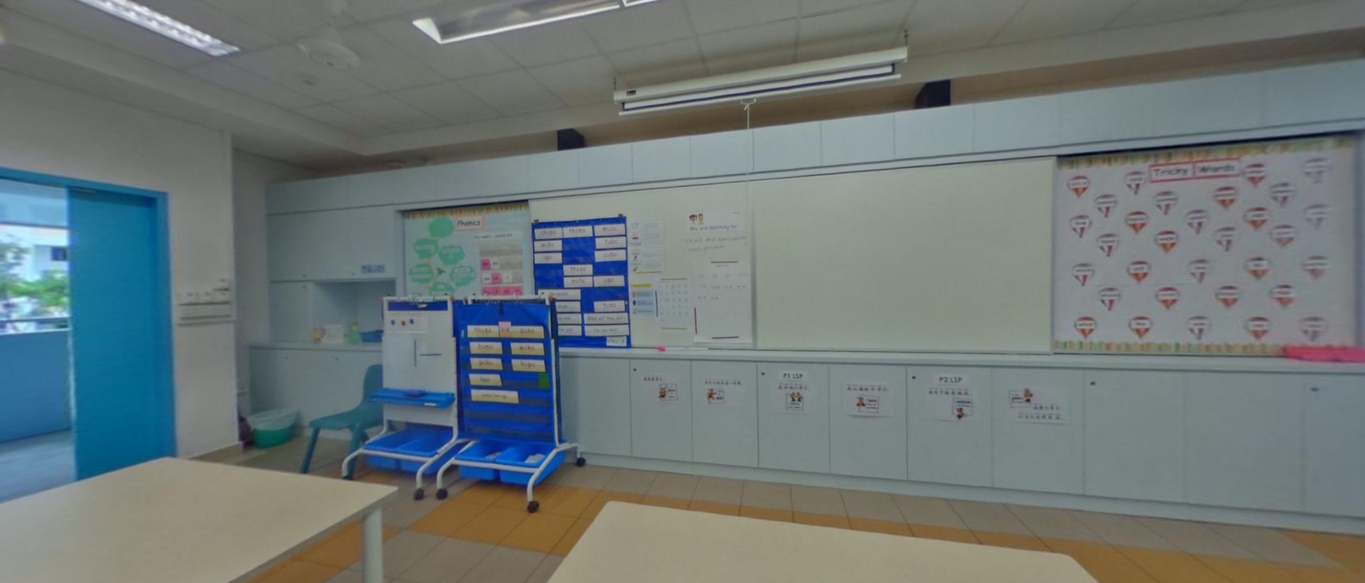 A classroom with whiteboards, colorful charts, blue storage carts, and student desks on a tiled floor.
