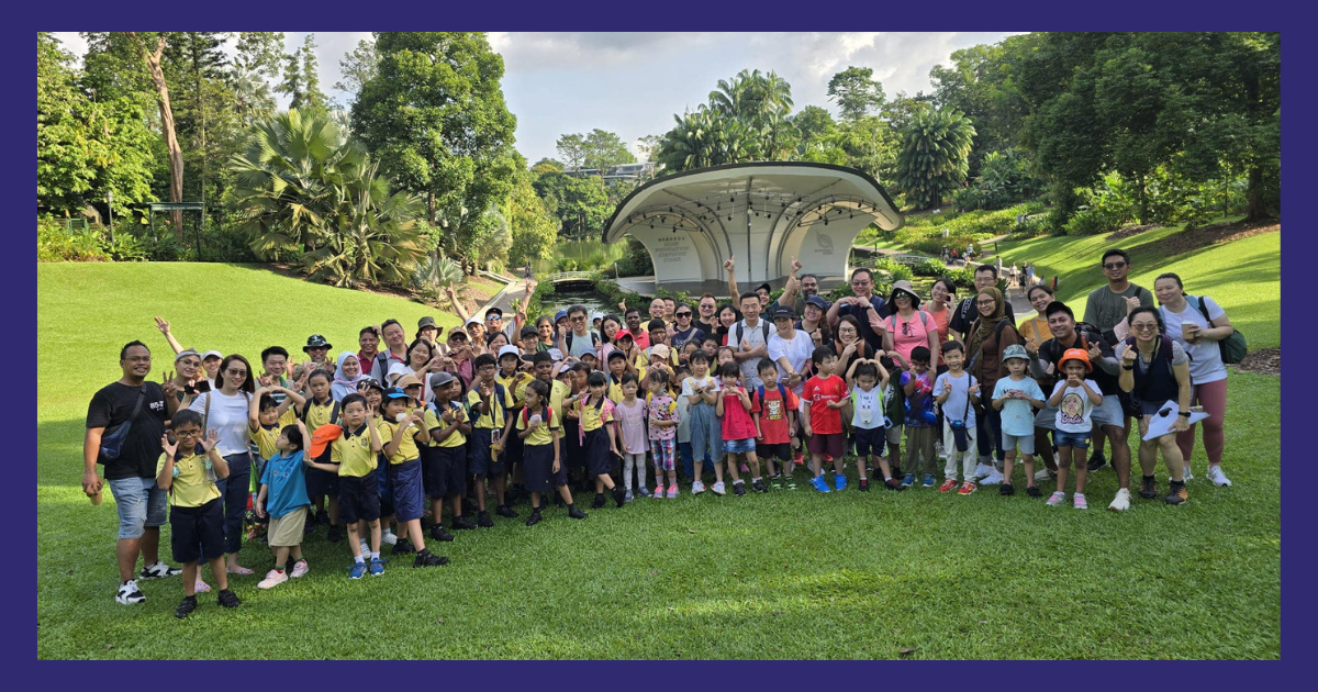 Large group of adults and young children in yellow uniforms posing on grass in a lush green park with an open-air stage.