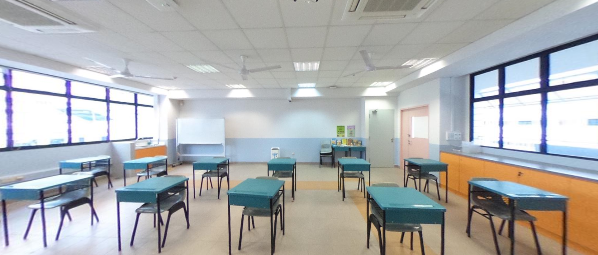 Empty classroom with teal student desks and chairs spaced apart, a whiteboard at the front, ceiling fans, and large windows.