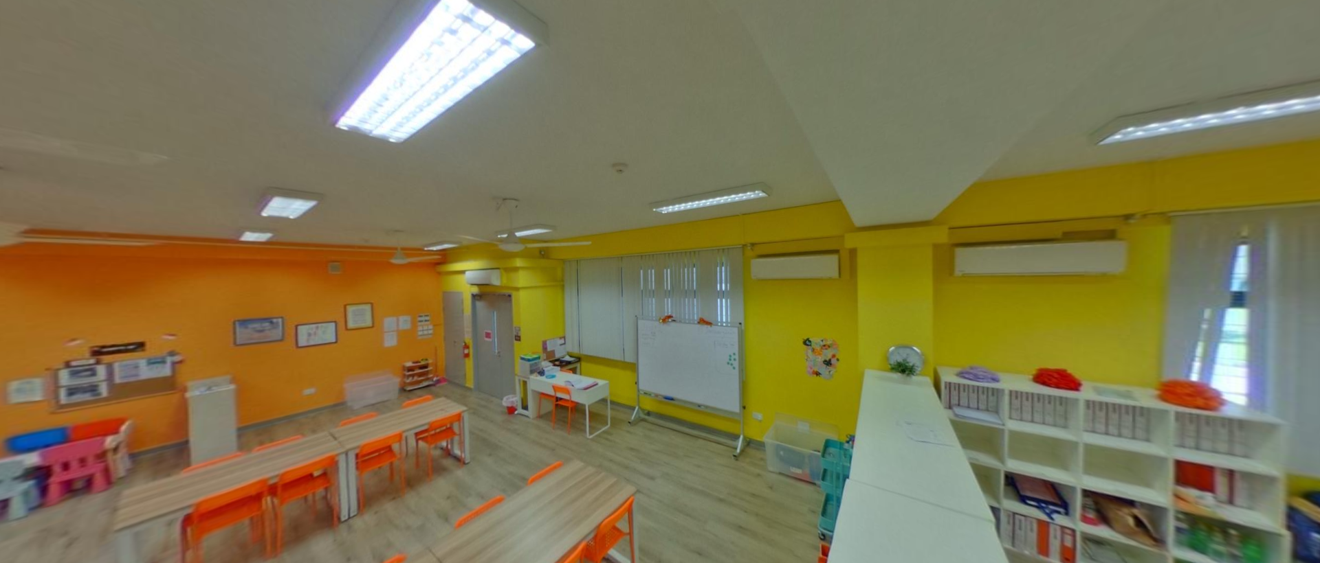 Bright classroom with orange and yellow walls, wooden desks, orange chairs, a whiteboard, and shelving units.