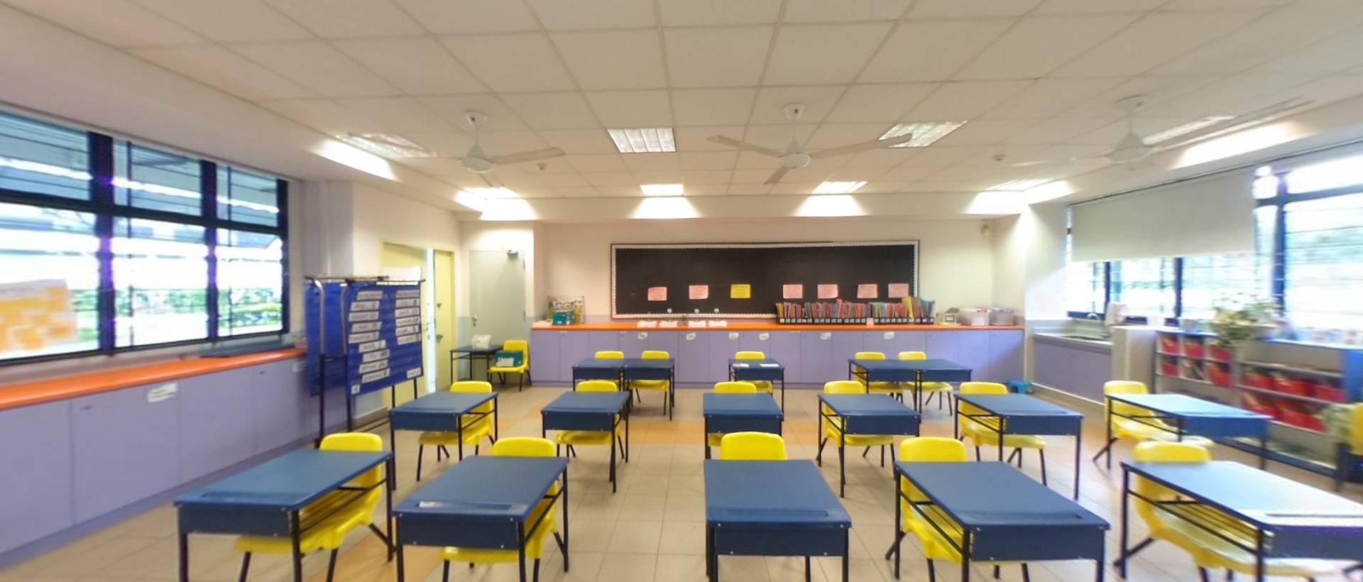 Empty classroom with rows of blue desks and yellow chairs facing a blackboard with pink notes.