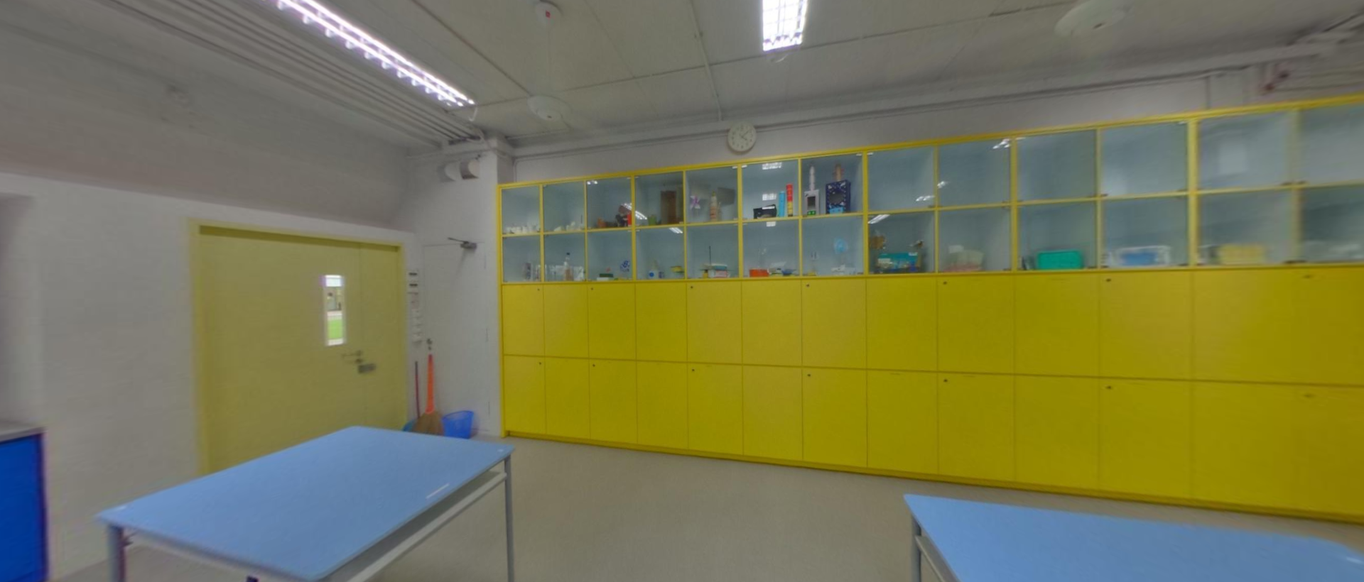 A bright room with yellow storage cabinets and glass shelves along the wall, blue tables, and a yellow door.