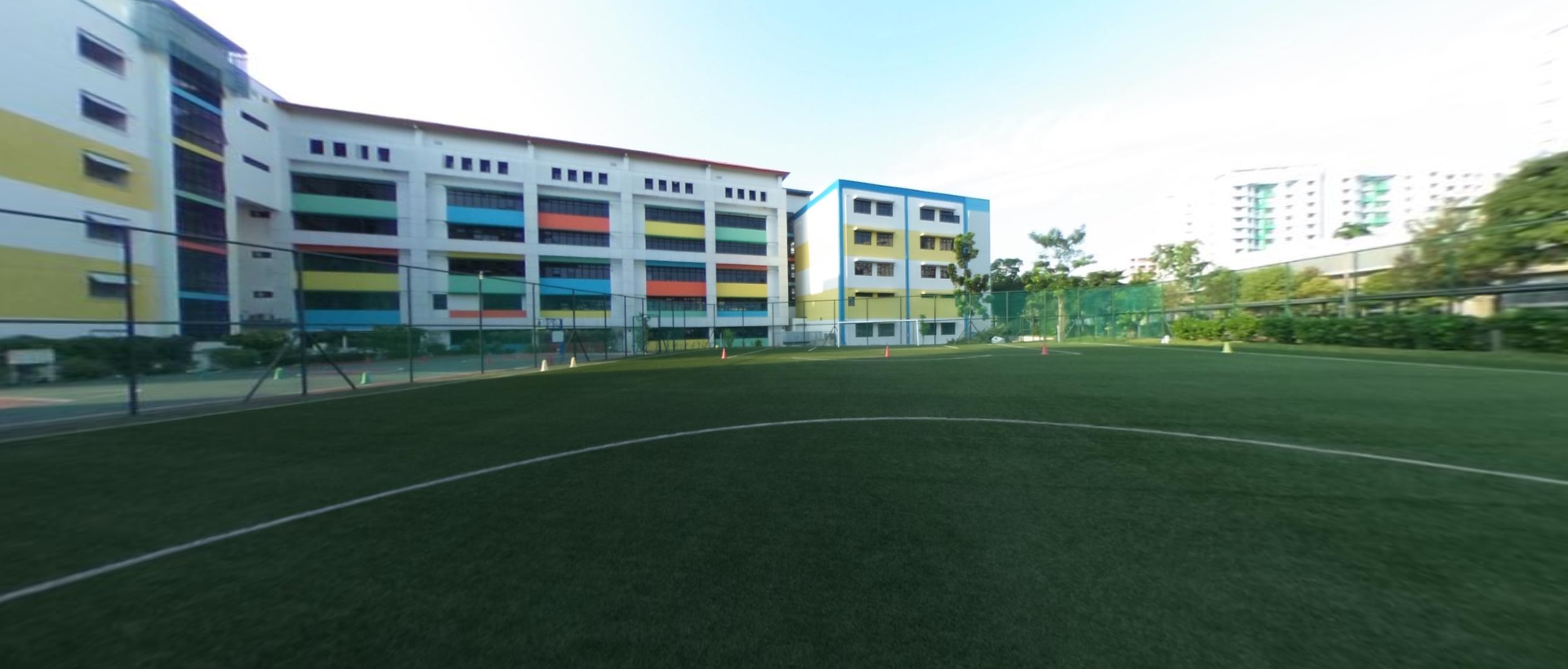 A green turf football field with orange cones, beside a colorful multi-story school building under a clear sky.