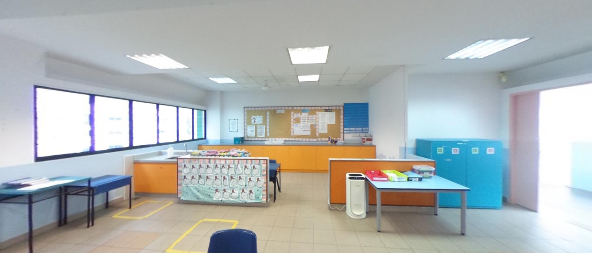A bright classroom with orange cabinets, blue tables, a bulletin board, and colorful storage lockers along the wall.