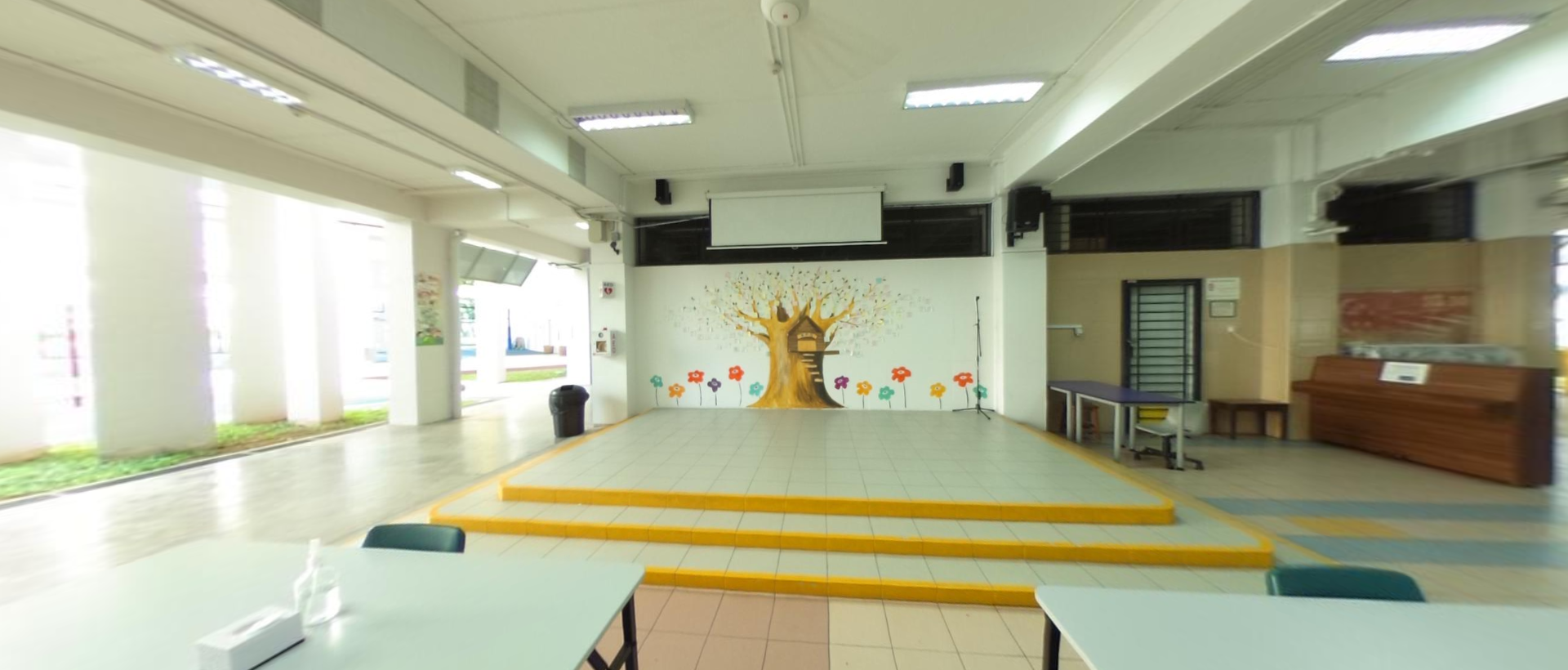 A school hall with a raised stage, projection screen, and a colorful tree mural with flowers on the back wall.
