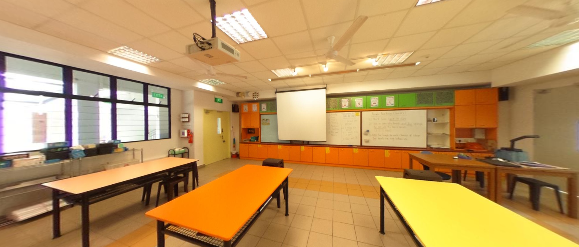 Empty classroom with orange and yellow tables, a whiteboard, projector screen, and orange cabinets along the wall.