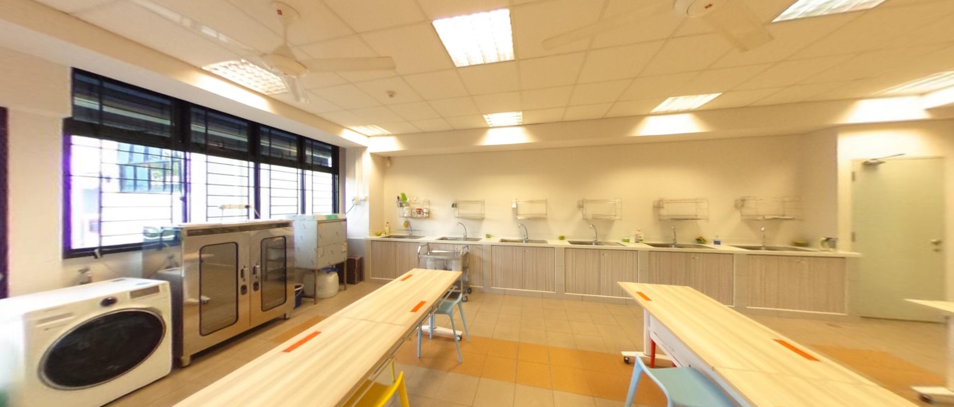 A bright classroom kitchen with long wooden tables, sinks along the wall, an oven, and a washing machine near the windows.