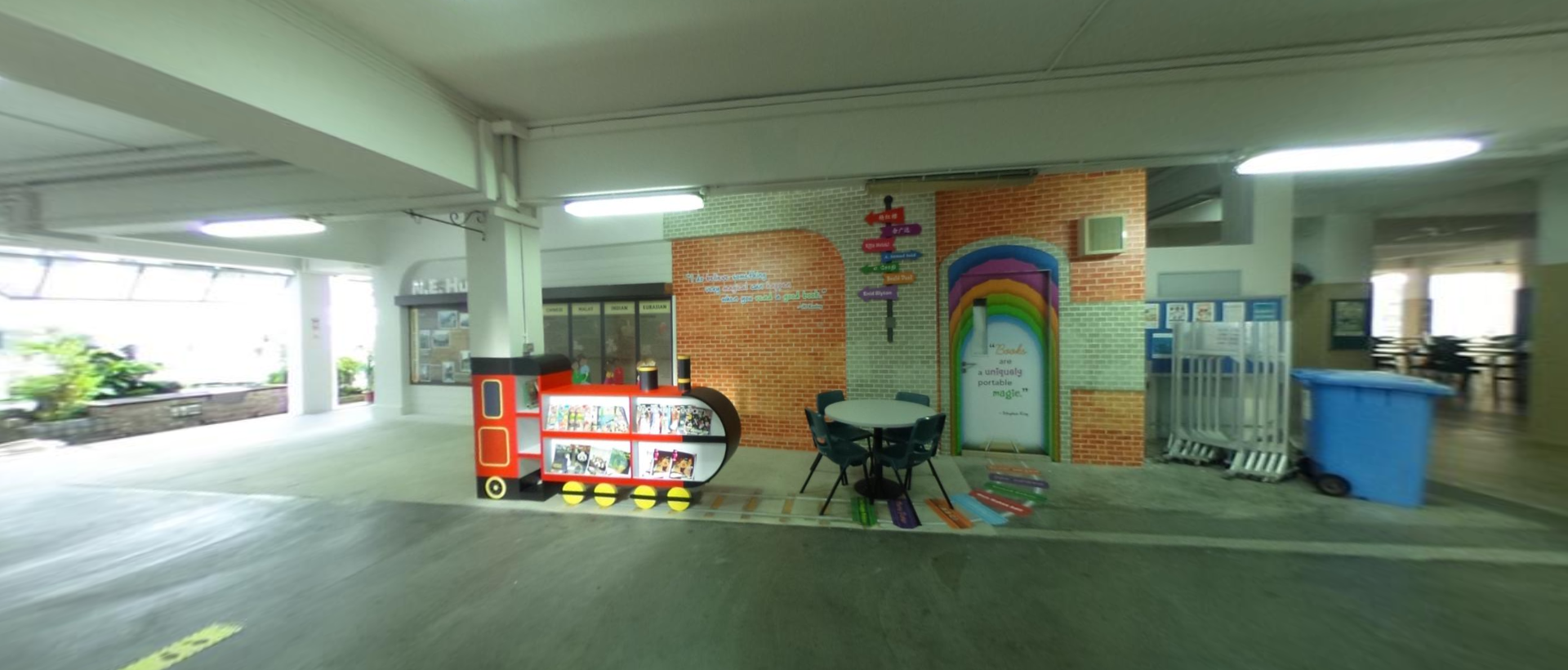 A red train-shaped bookshelf and small table with chairs beside a colorful rainbow mural on a brick wall indoors.