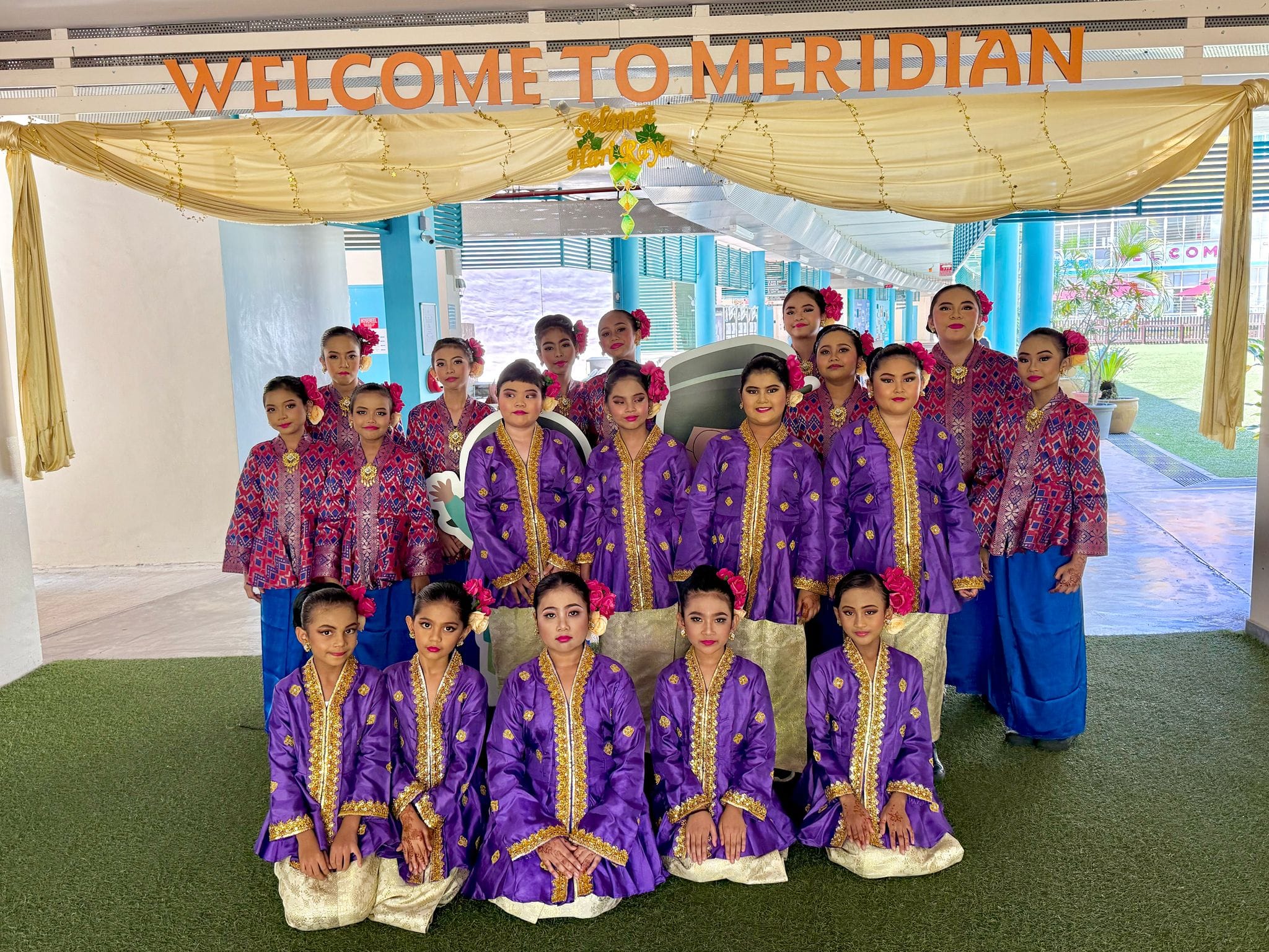Group in purple and blue traditional outfits posing under a WELCOME TO MERIDIAN banner and gold drape.