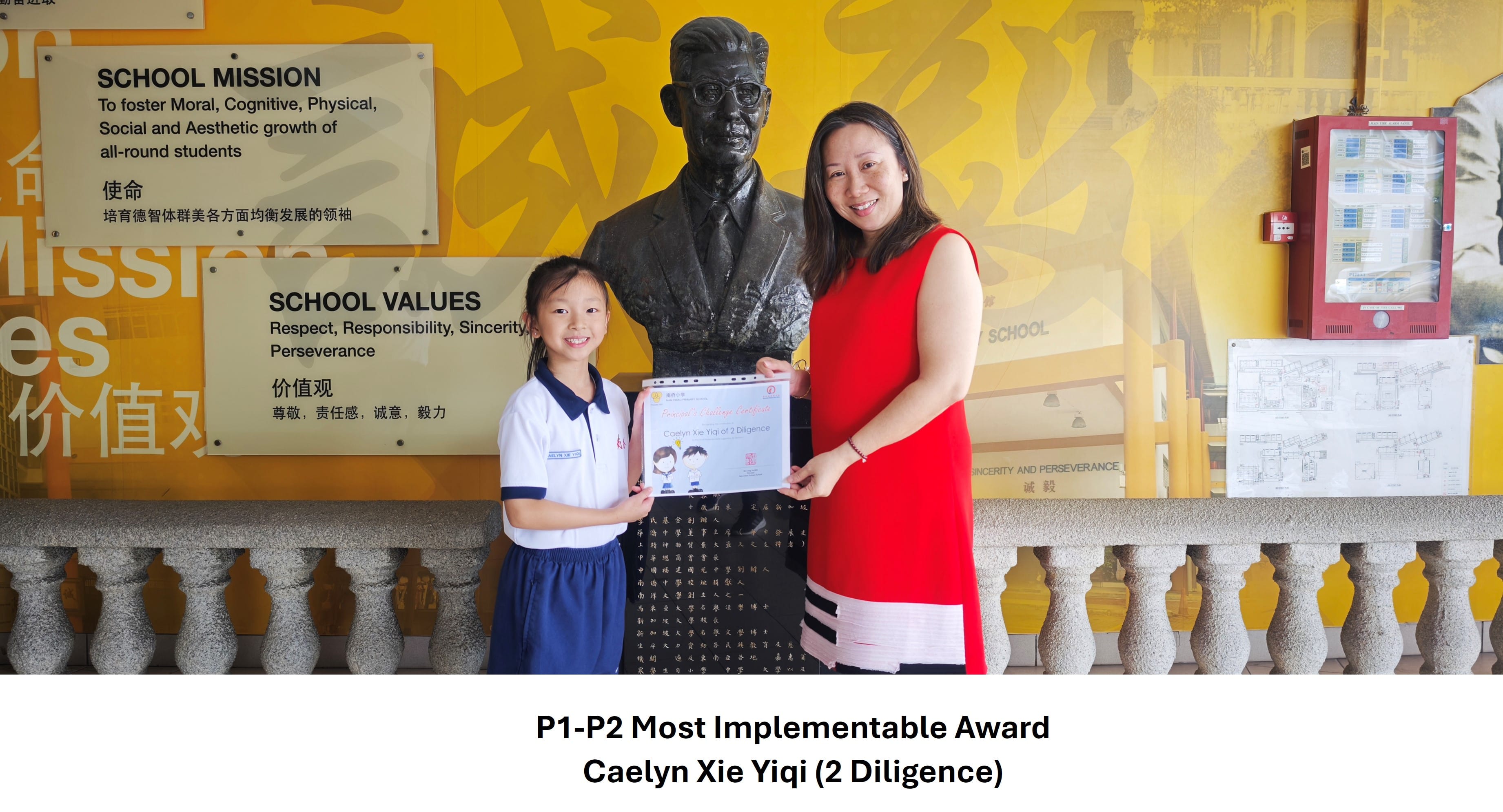 A young girl and a woman in red hold a certificate together in front of a bronze bust and school mission wall.