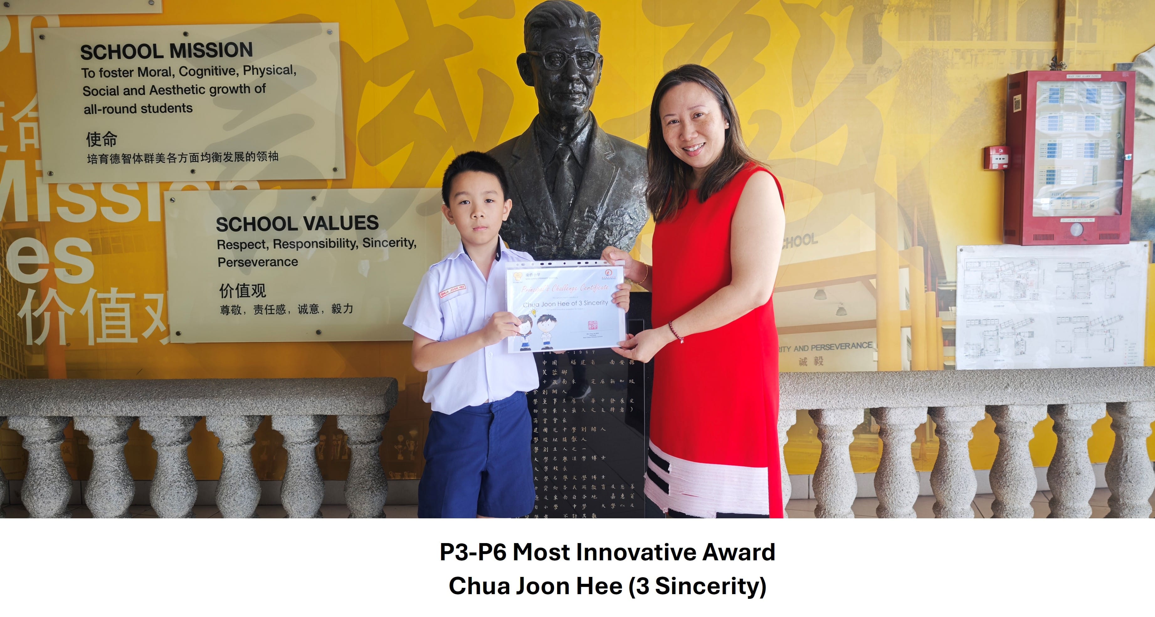 A young boy in school uniform and a woman in red hold a certificate before a bust statue and school mission board.