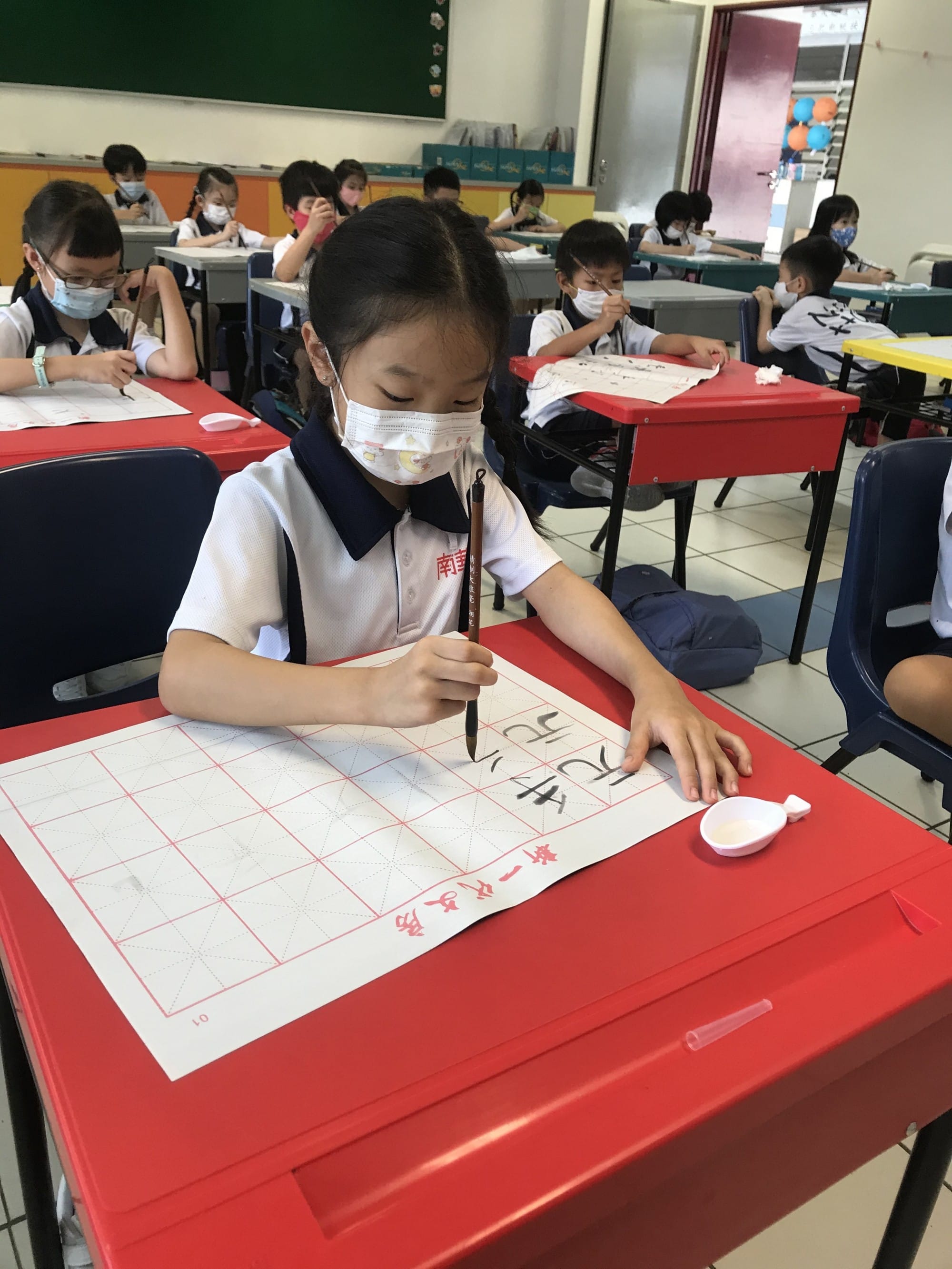 Young girl in uniform and mask practices Chinese calligraphy with a brush at a red desk in a classroom.