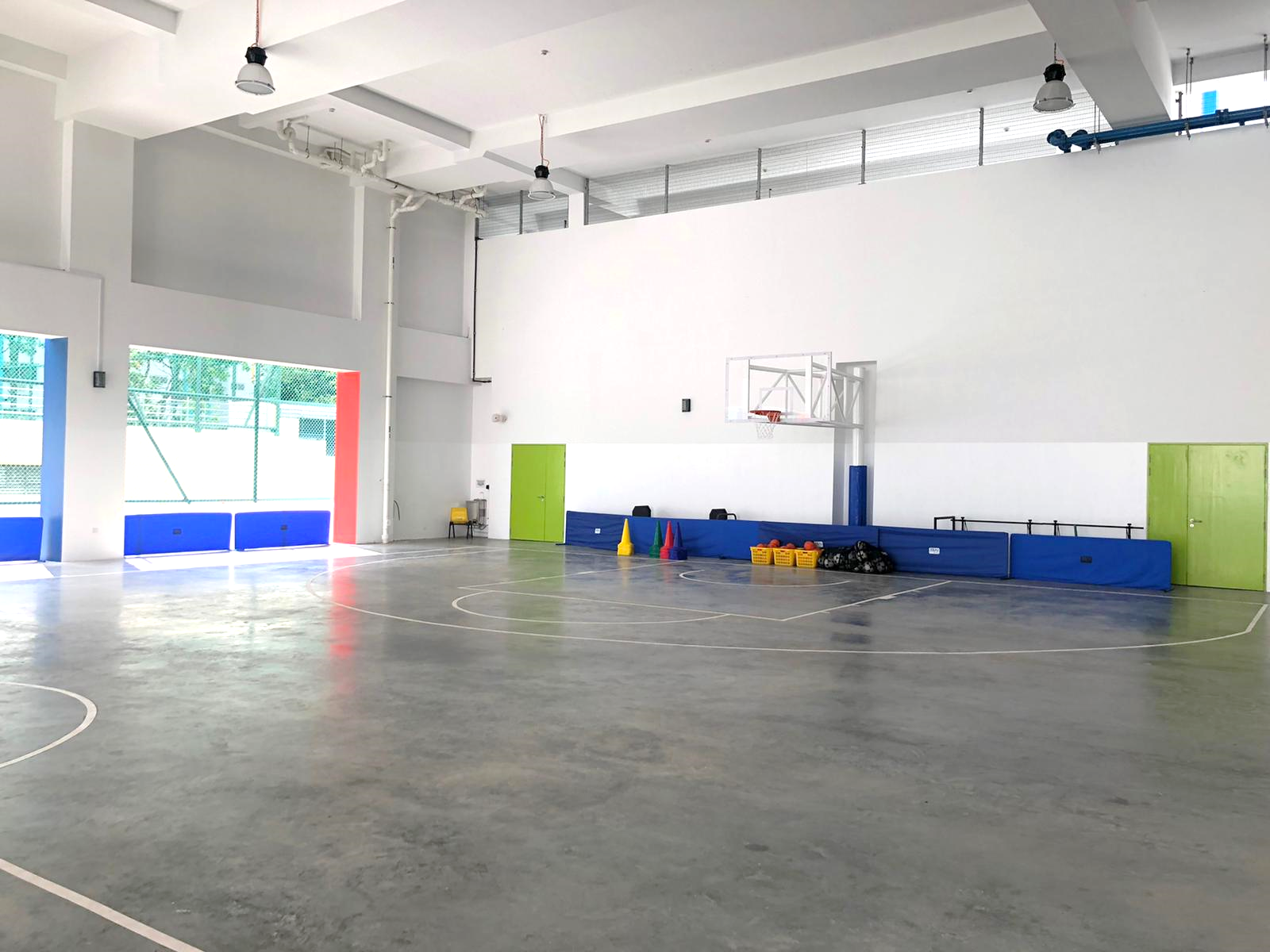 Indoor basketball court with gray floor, a hoop on a white wall, blue padding, and colorful cones and balls along the side.