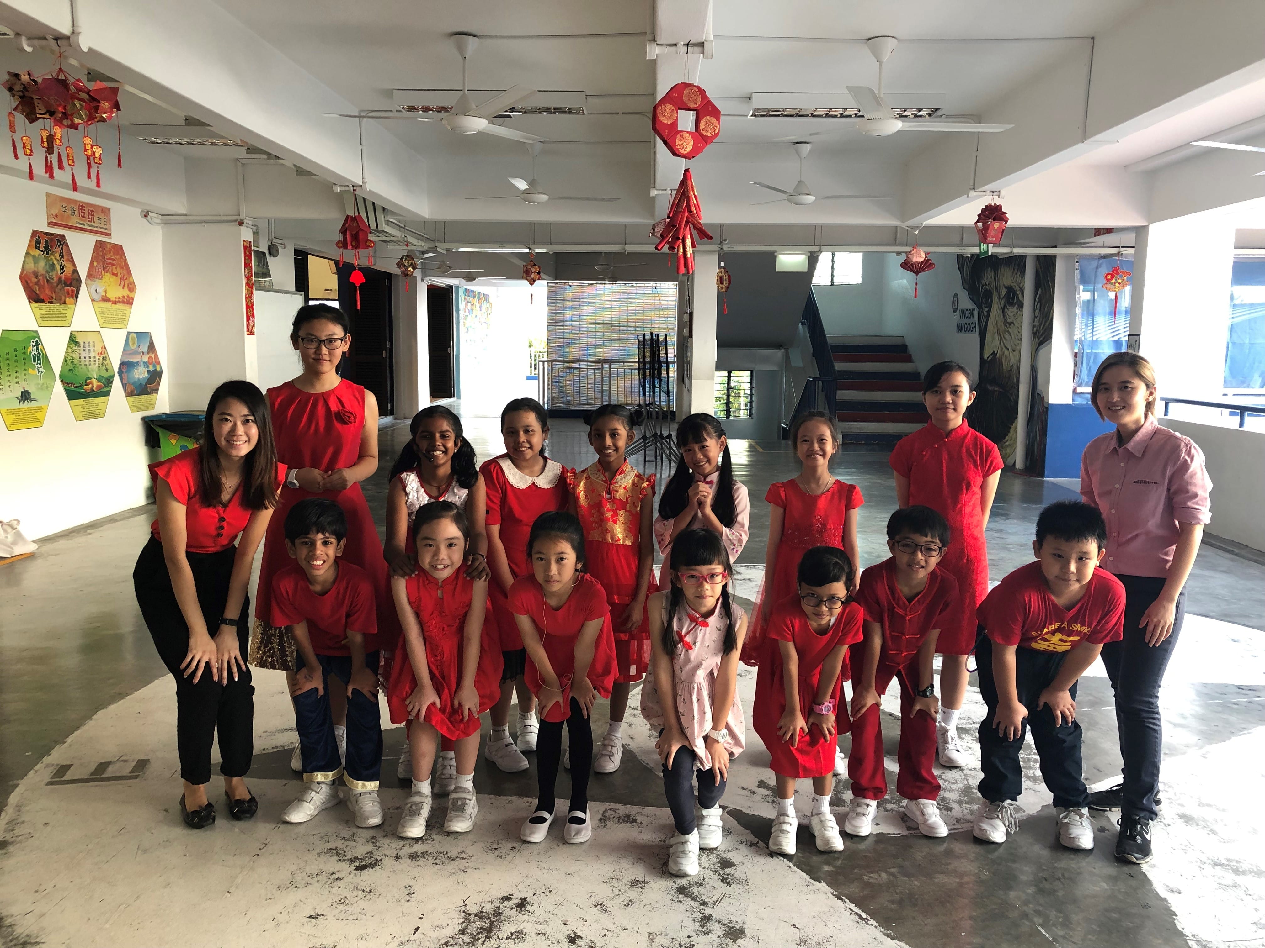 A group of children and adults wearing red outfits pose indoors decorated with Chinese New Year ornaments.