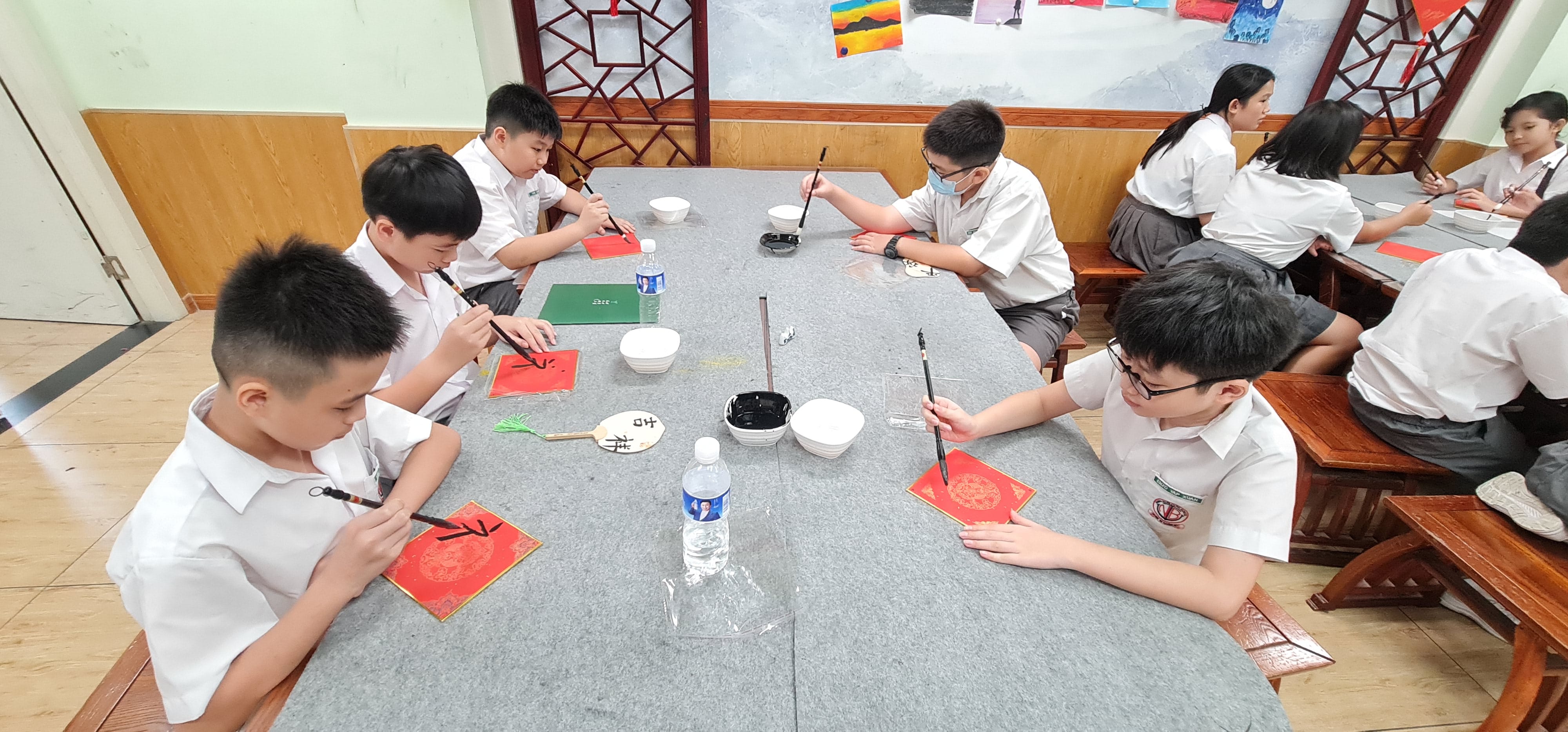 Students in white uniforms practice Chinese calligraphy with brushes on red paper at a classroom table.