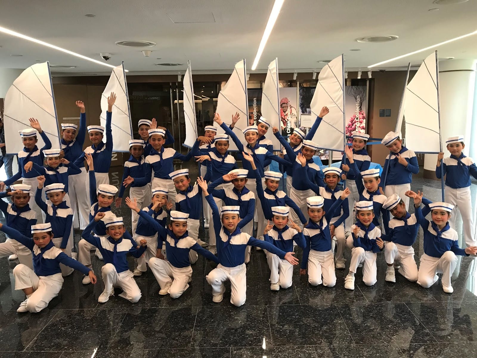 A large group of children in blue and white sailor costumes pose with white sail props in an indoor venue.