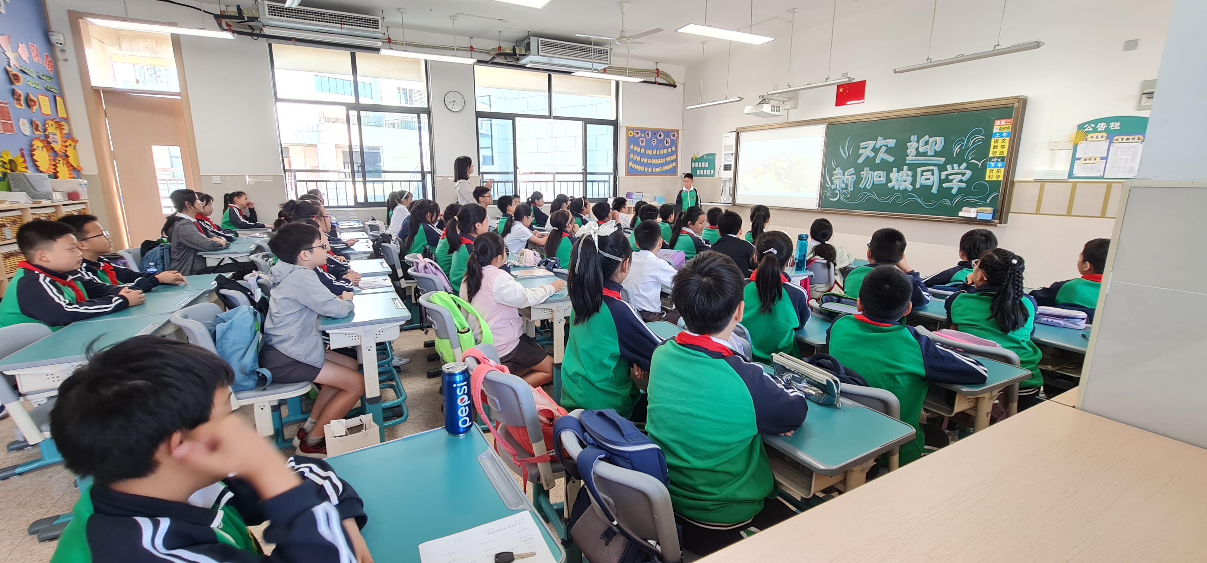 Students in green uniforms sit at desks facing a chalkboard with Chinese writing in a bright classroom.