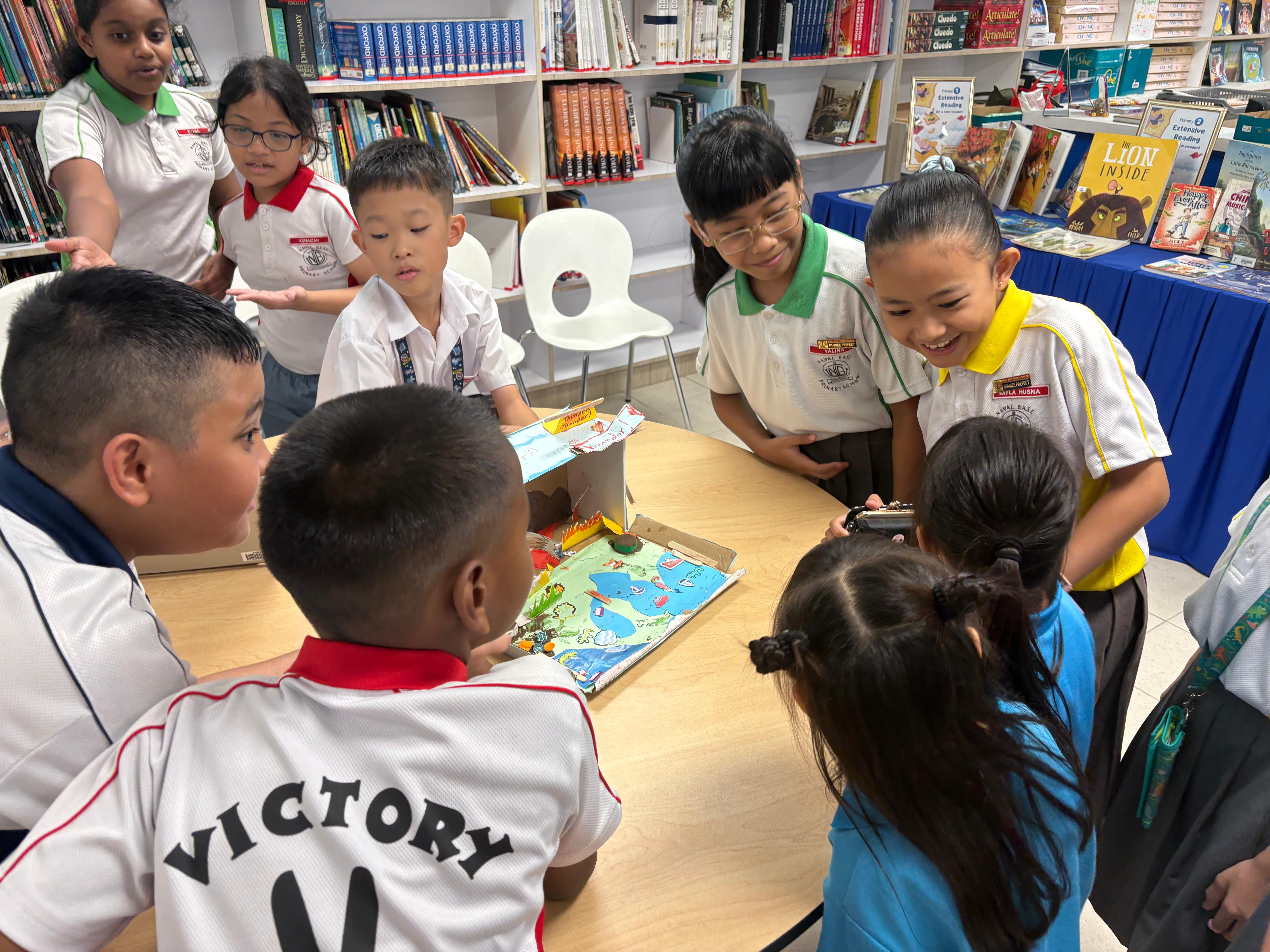 Several school children in uniforms gather around a table in a library, looking at a colorful diorama project.