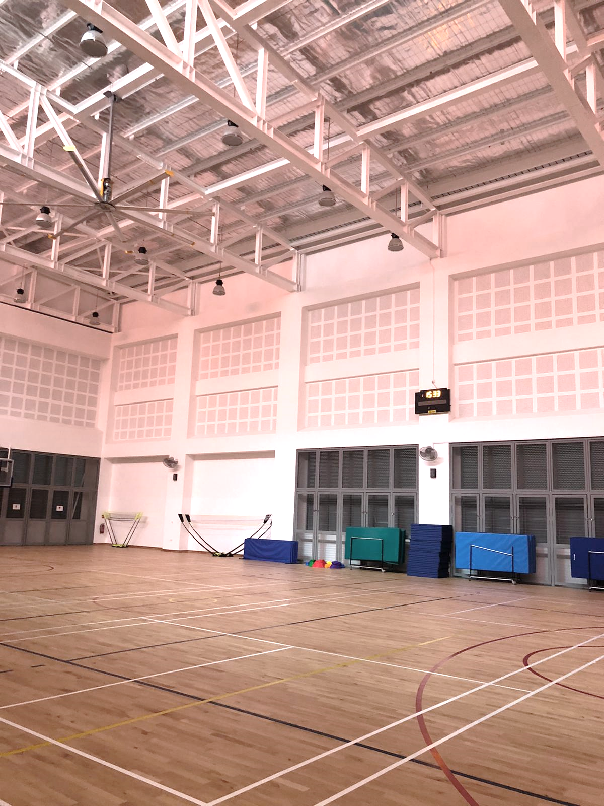 Empty indoor sports hall with wooden floor, court lines, high white walls, steel roof trusses, and blue gym mats along the wall.