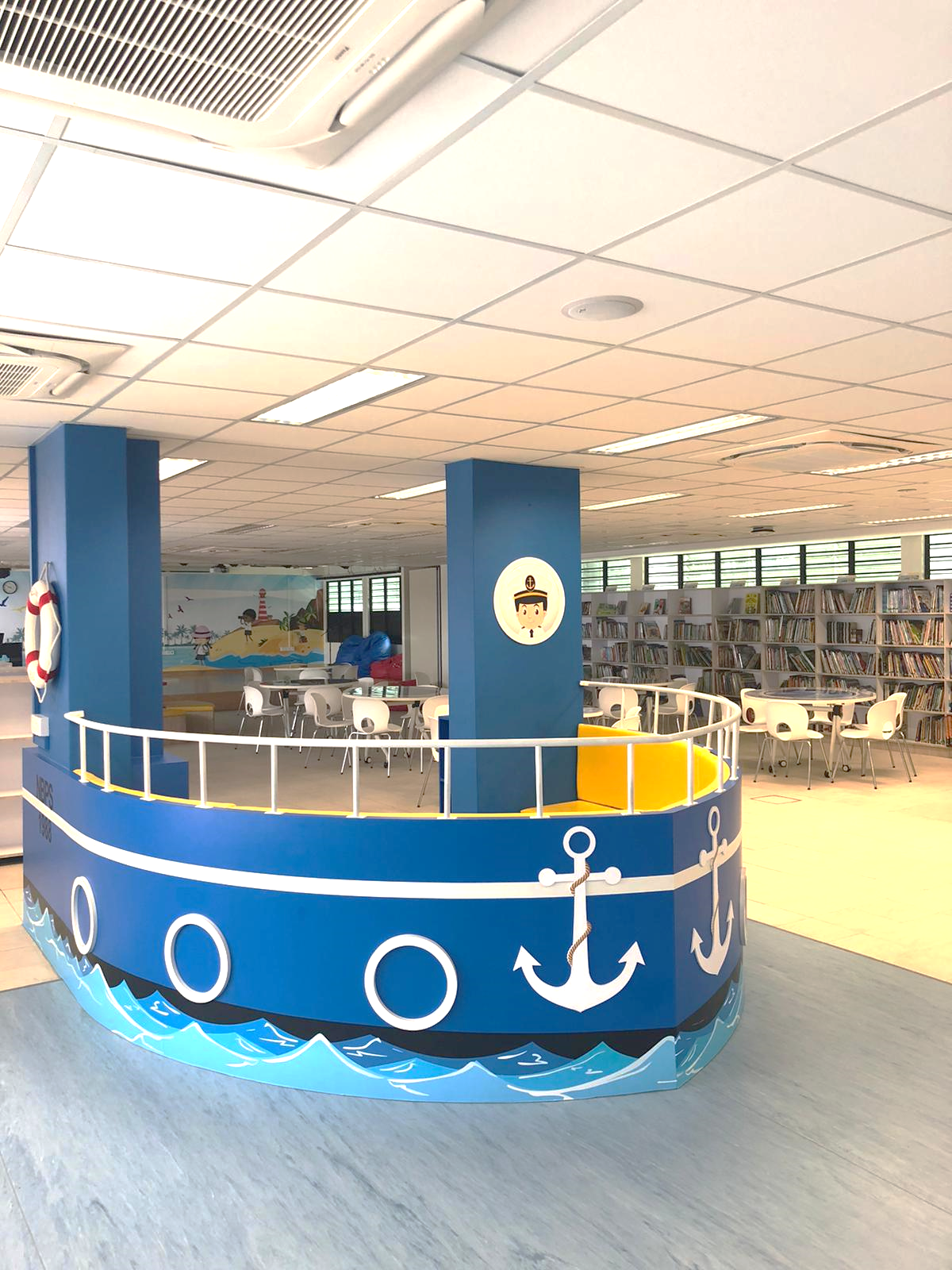 A children's library with a blue boat-shaped reading nook decorated with anchors, surrounded by bookshelves and seating areas.