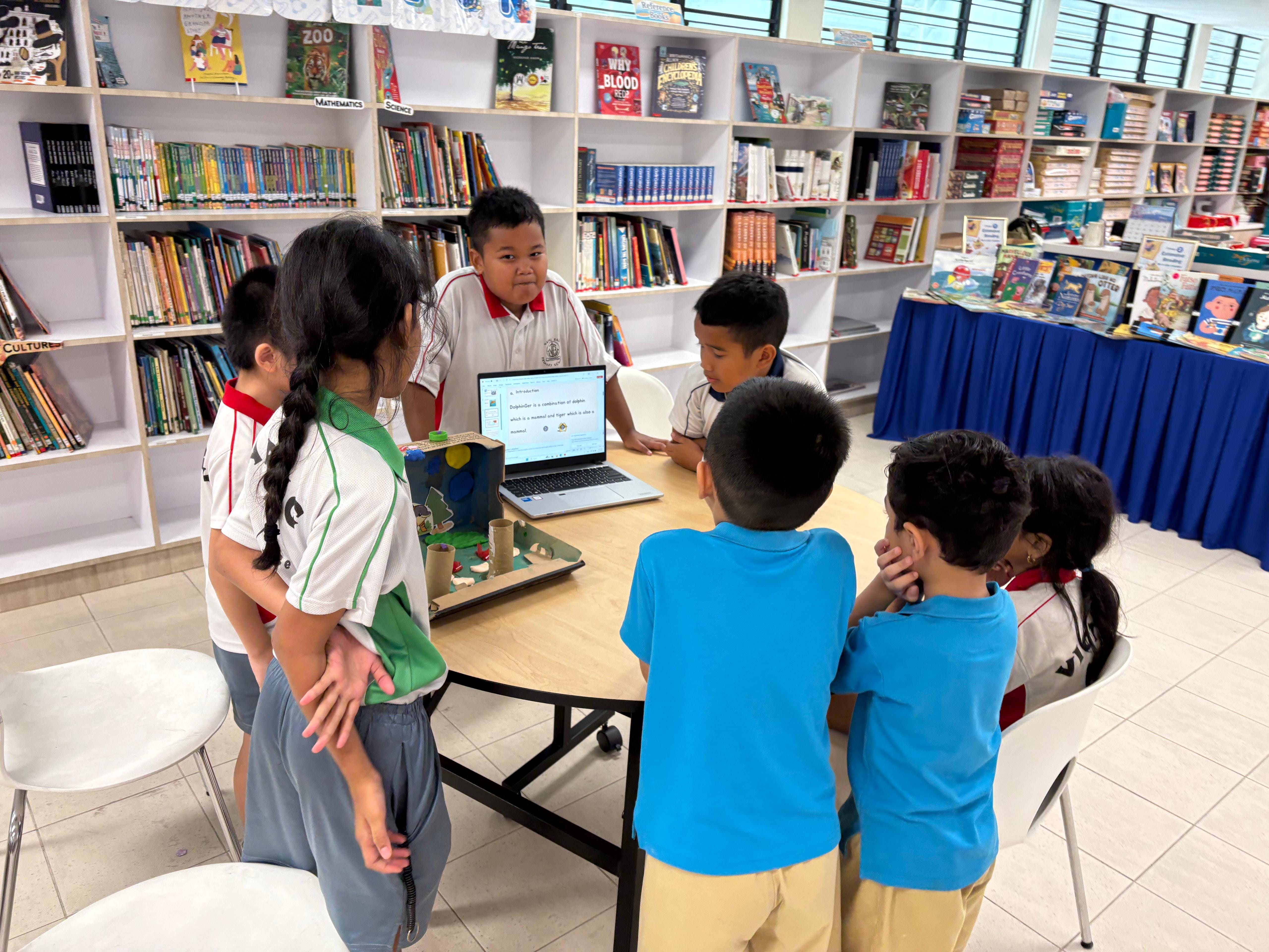 Six children gather around a round table in a library, viewing a laptop and a small handmade model project.