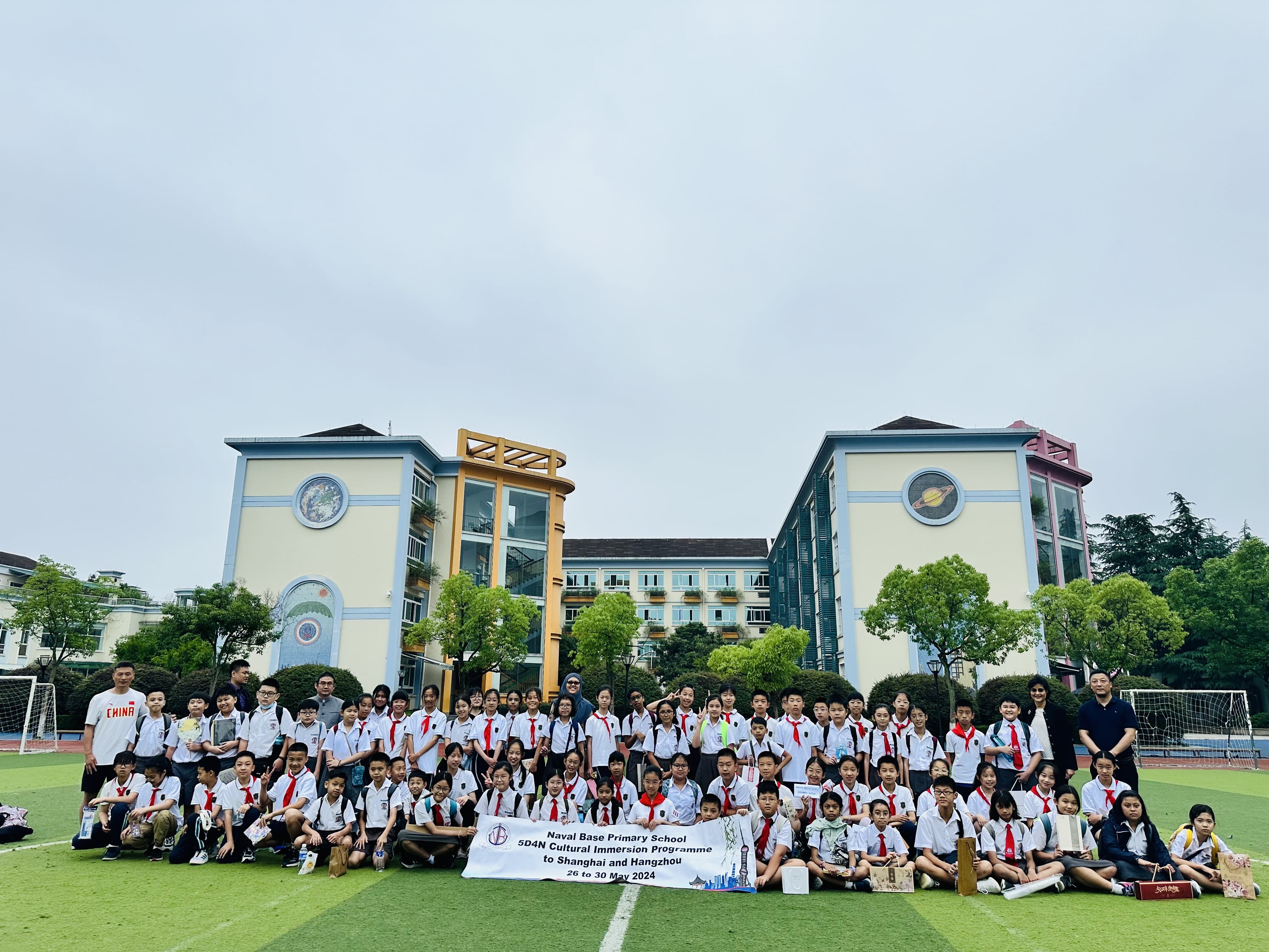 Large group of students in white uniforms with red ties posing outdoors in front of a school building holding a banner.