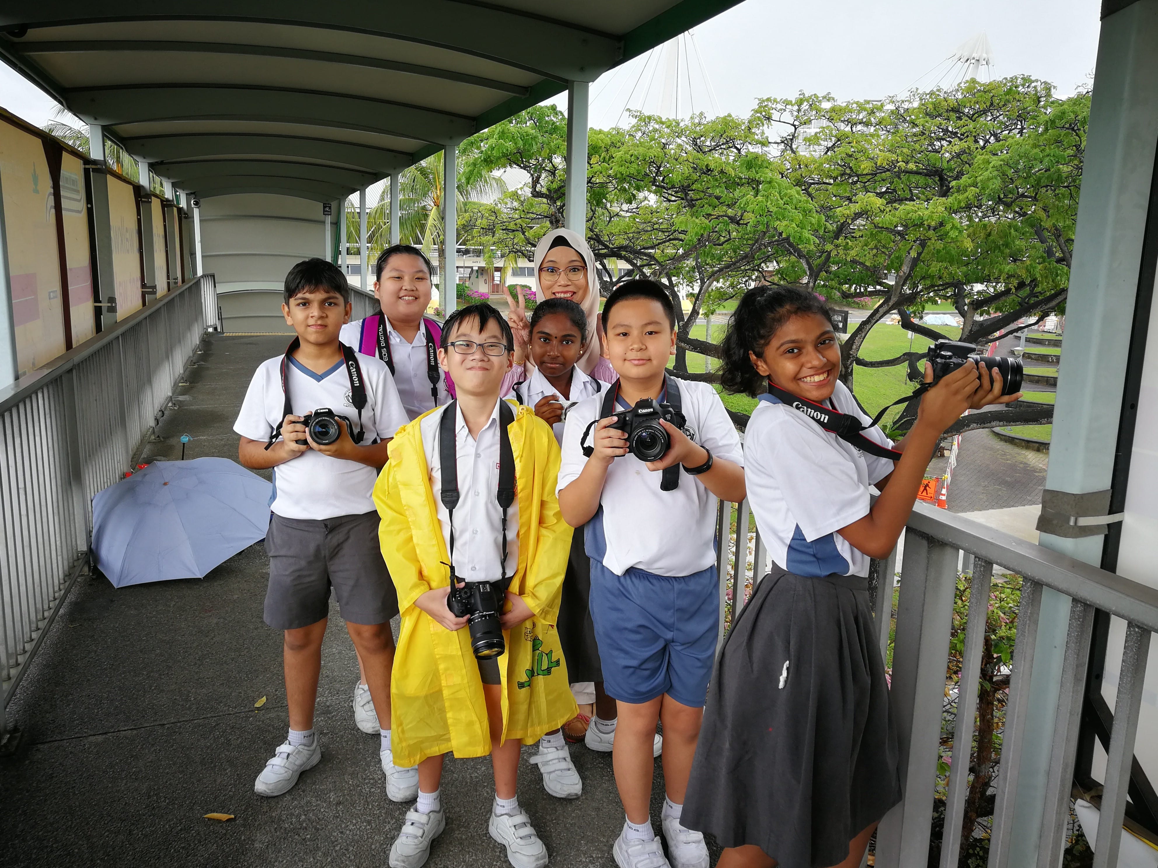 Seven school children in uniforms holding Canon cameras stand smiling on a covered outdoor walkway.