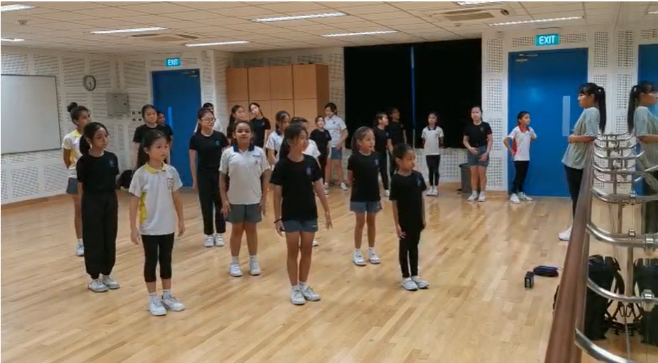 A group of children stand in rows on a wooden floor in a dance or rehearsal studio with blue doors.