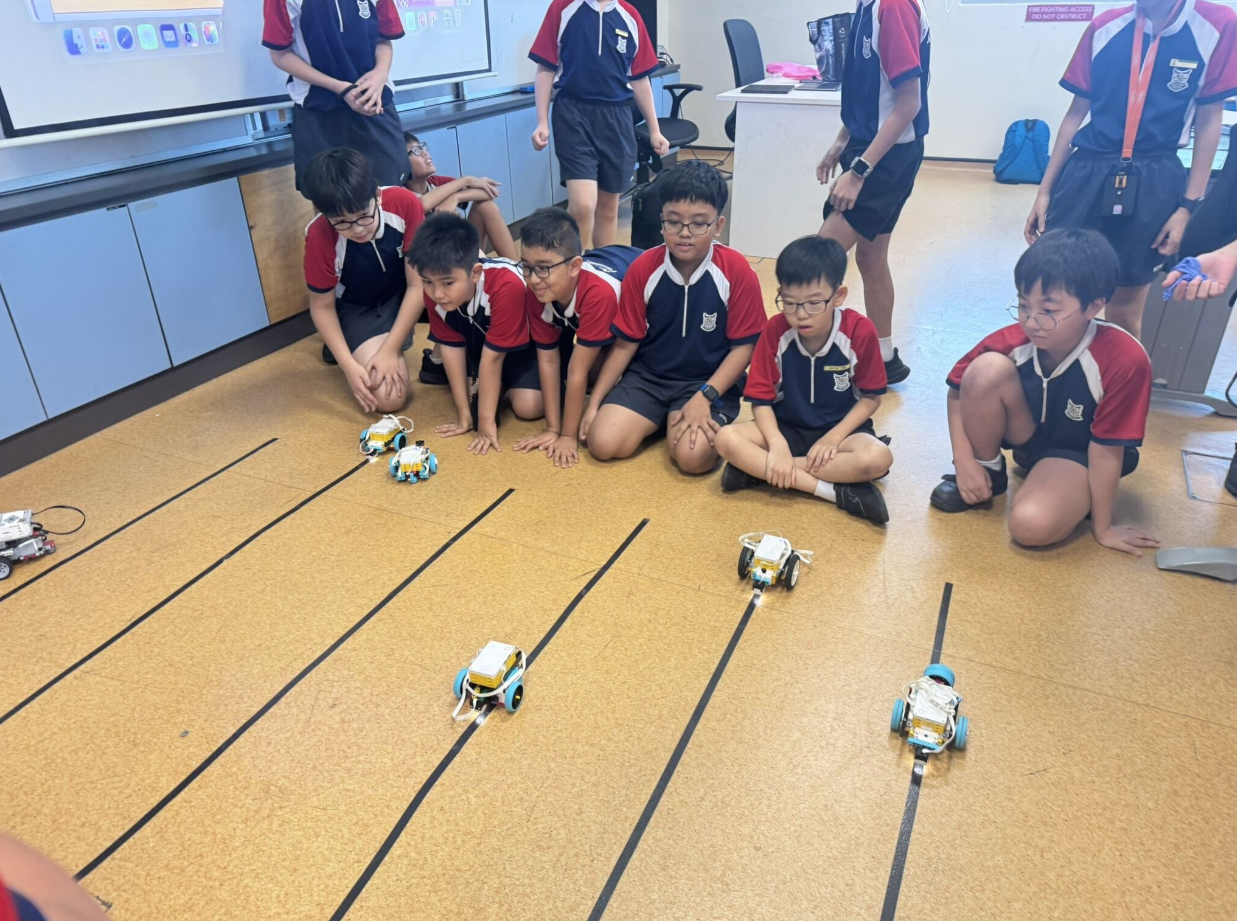 Students in school uniforms sit on a floor watching small robots race along black tape track lanes.