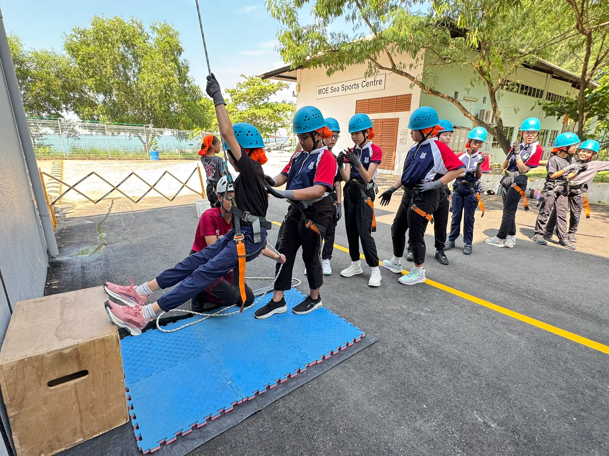 Students in blue helmets and harnesses practice rappelling at the MOE Sea Sports Centre outdoors.