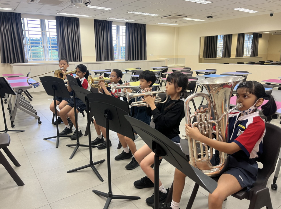 Six young students in school uniforms sit in a row playing brass instruments with music stands in a classroom.
