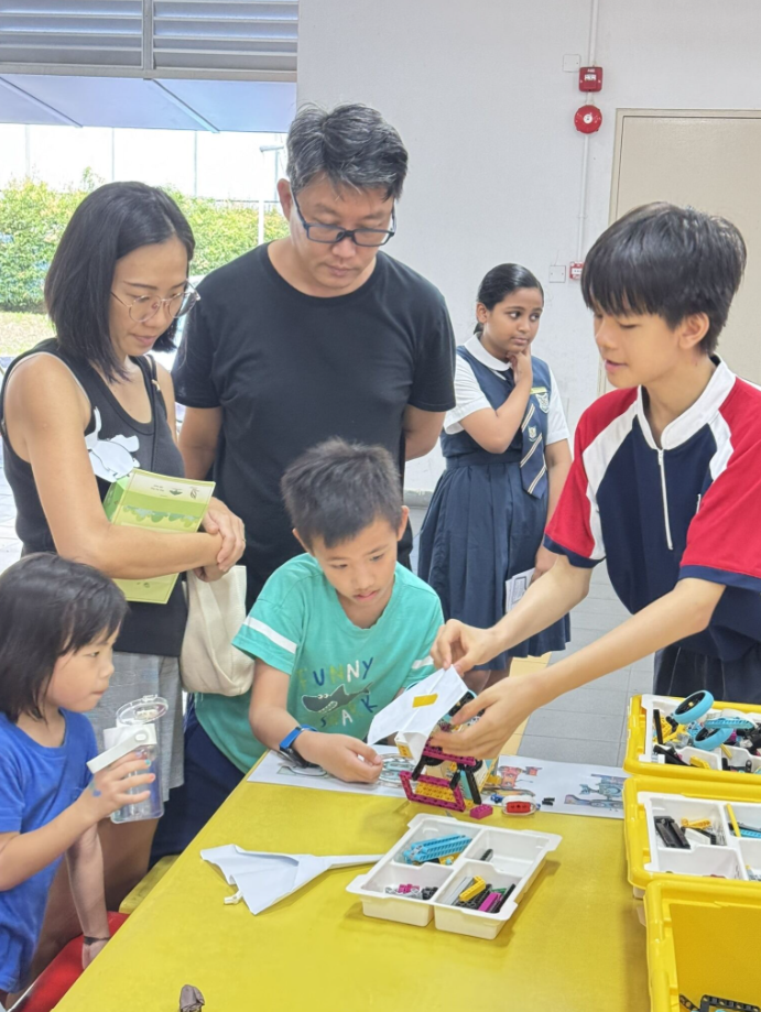 Adults and children gather around a yellow table building with LEGO bricks and sorted parts in trays.