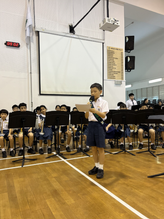 A young student speaks into a microphone holding paper, with seated students and music stands behind in a school hall.
