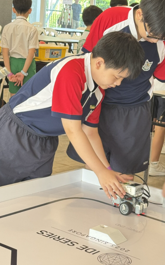 A student in school uniform leans over a table, placing a small LEGO robot on a competition mat labeled IDE Series Singapore.