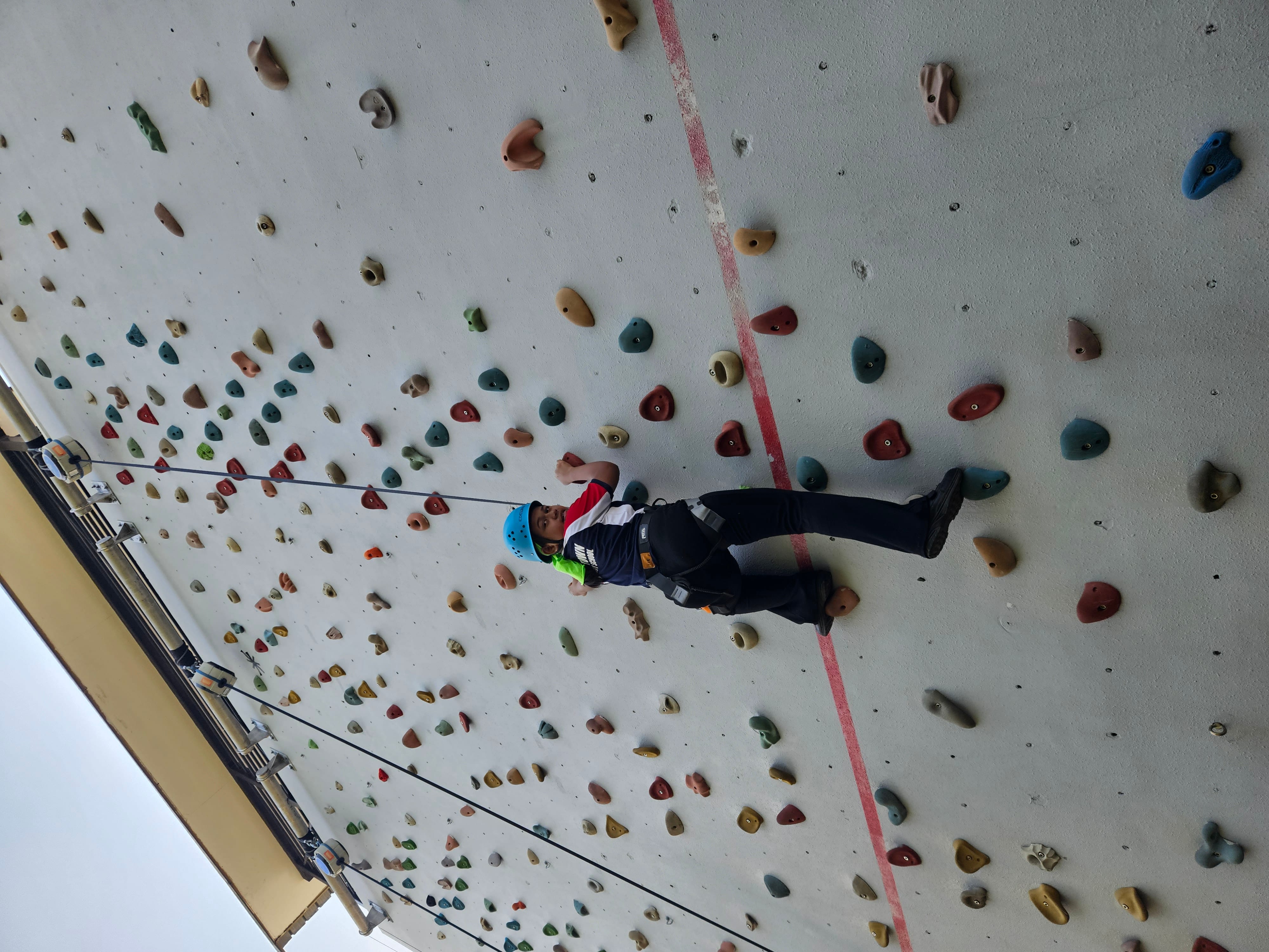 A child wearing a blue helmet and harness climbs a tall outdoor rock wall with colorful holds.