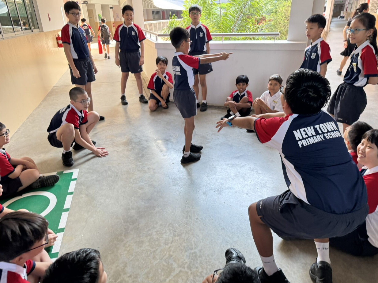 Students in New Town Primary School uniforms sit and stand in a circle outdoors during a group activity.