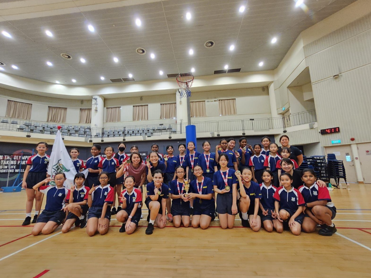 A large group of young girls in blue and red uniforms pose with trophies and medals on an indoor basketball court.