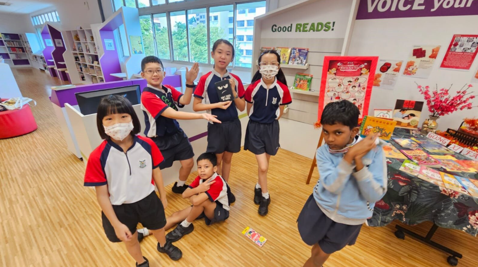 Five school children in uniforms pose and smile in a colorful library with a Good Reads sign and book displays.