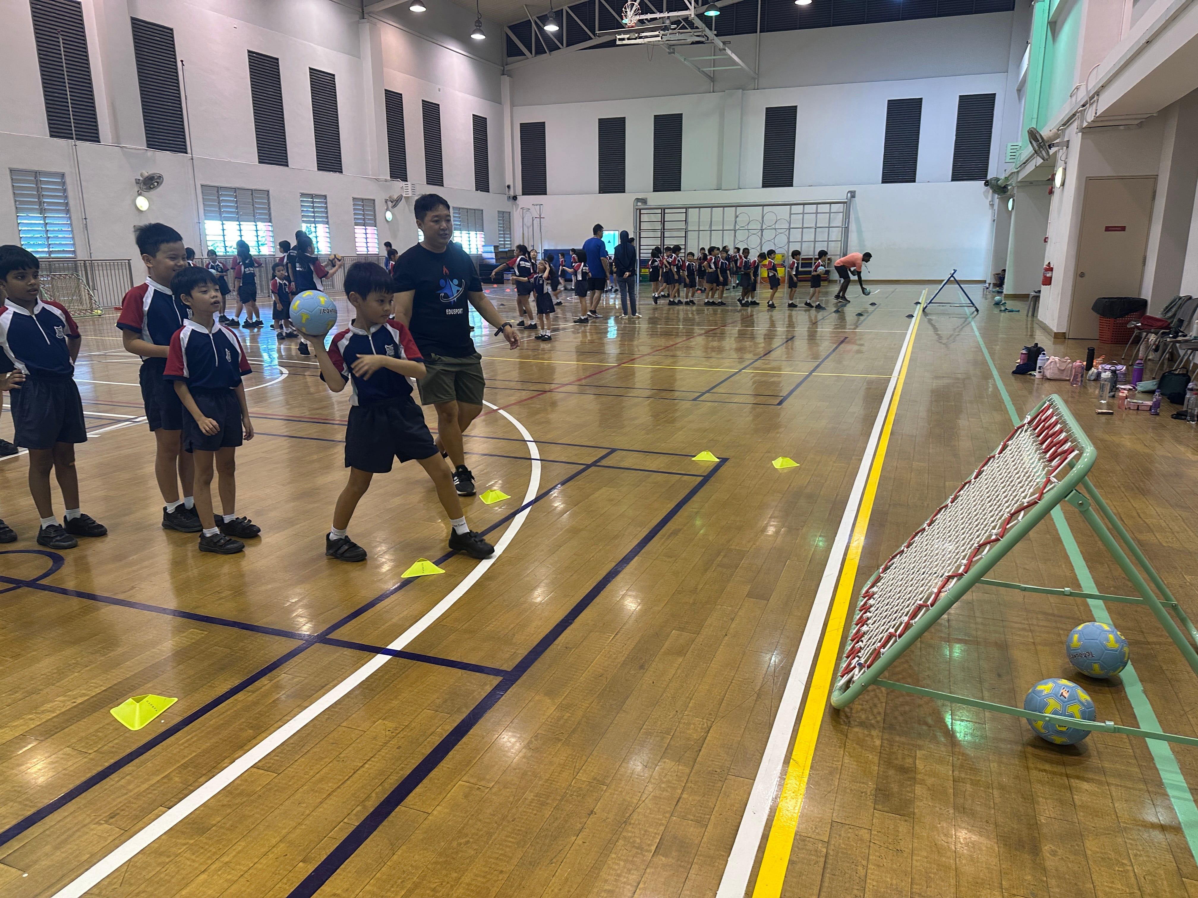 Children in school uniforms practice ball skills in an indoor gym, guided by an adult coach near a small goal net.