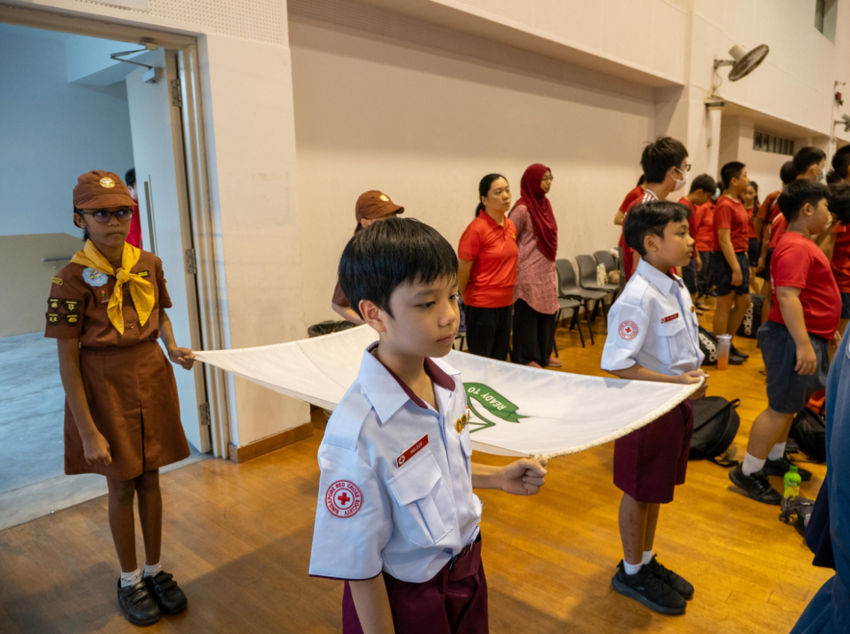 Children in Red Cross and scout uniforms carry a white banner in an indoor hall with adults watching nearby.