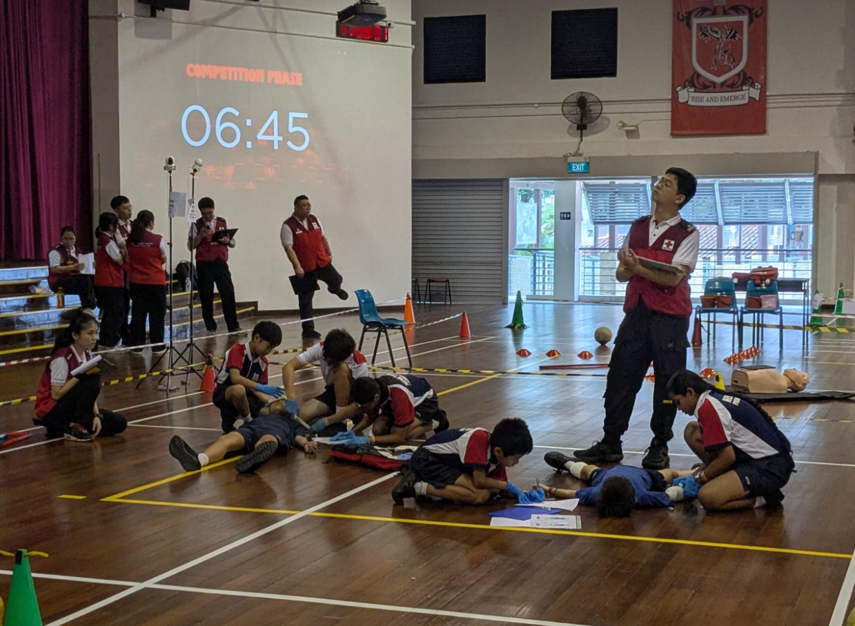 Students in red vests practice first aid on mannequins in a hall during a timed competition phase showing 06:45.