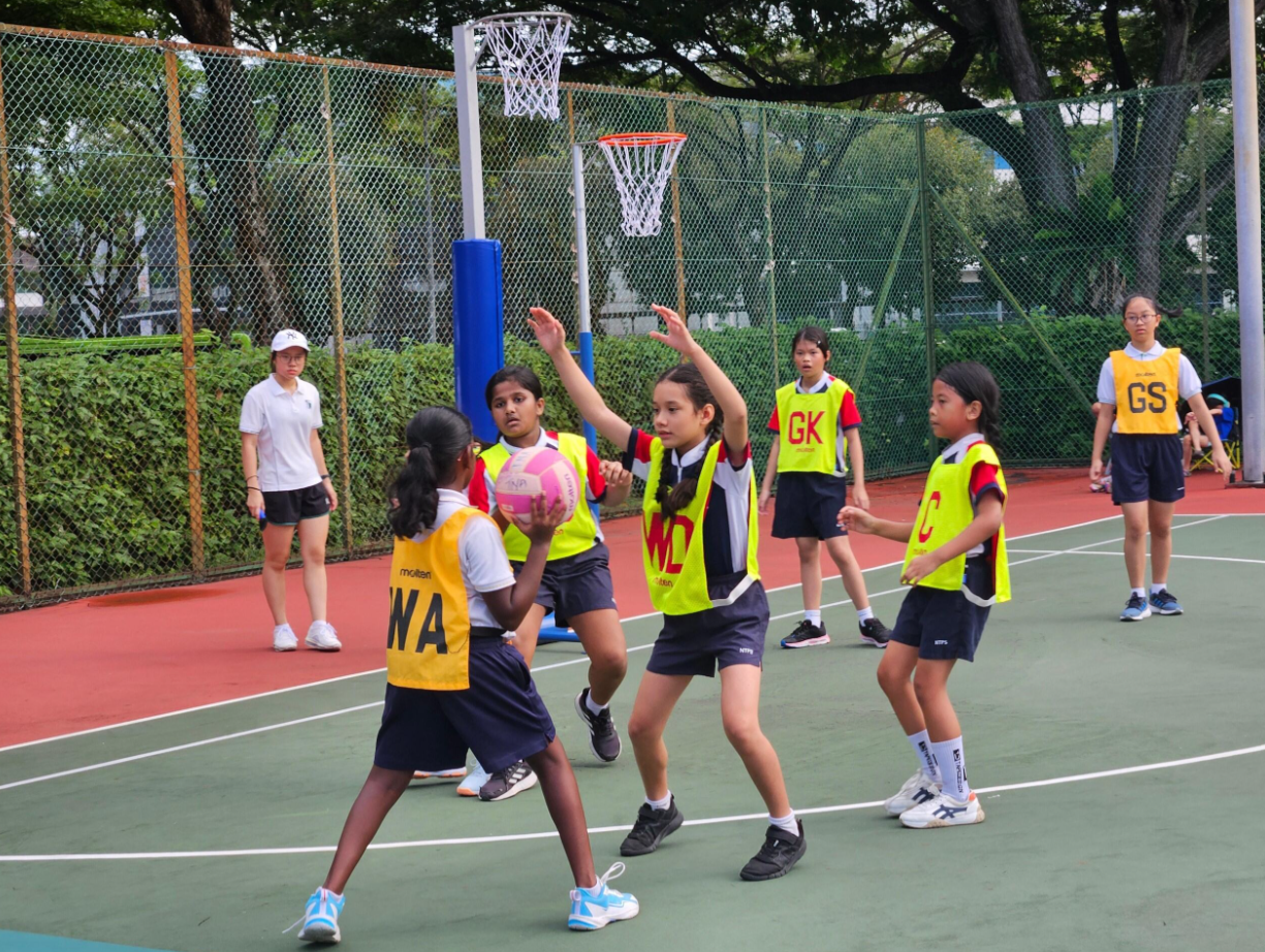 Girls in yellow and blue bibs playing netball on an outdoor court, one holding a pink ball near the hoop.