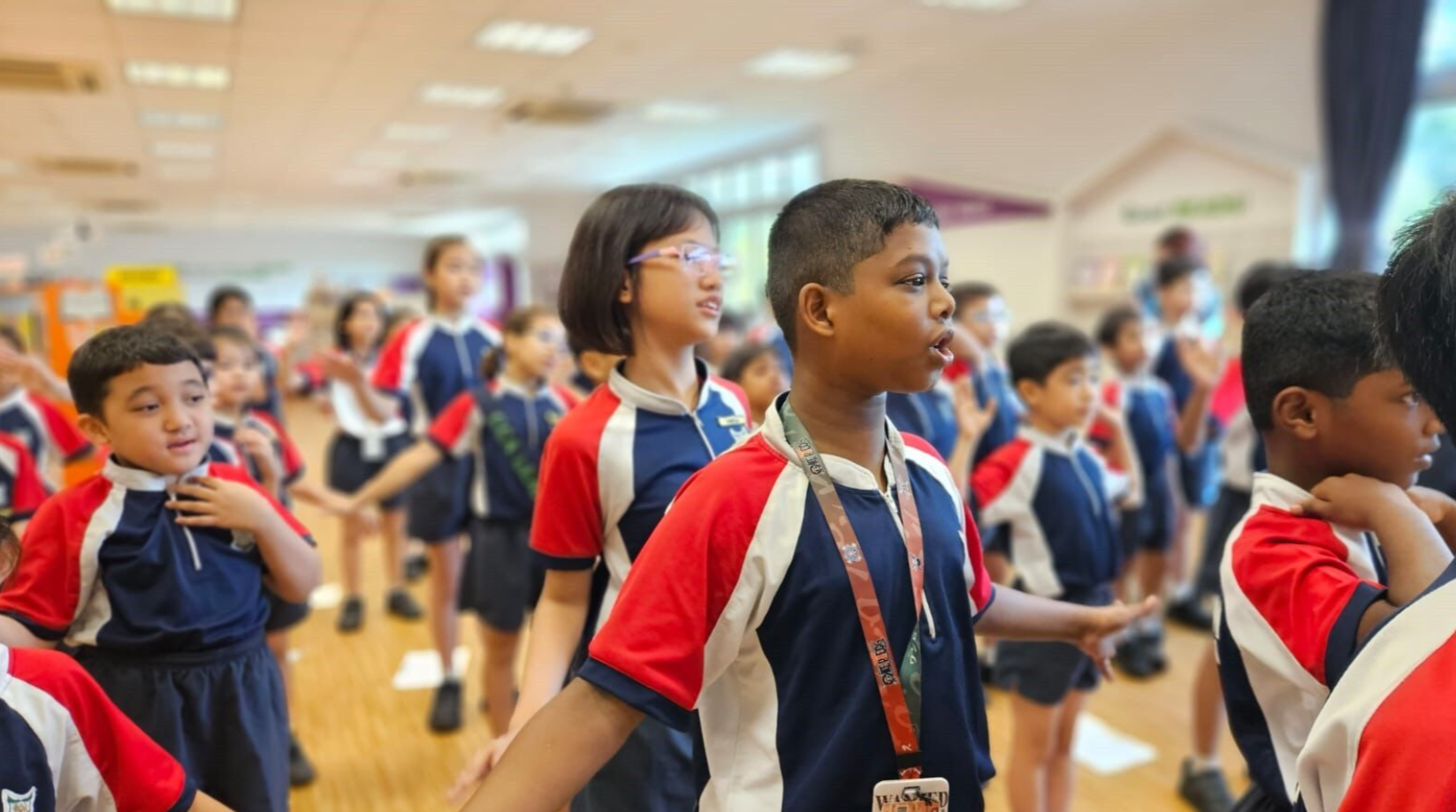 A group of school children in red, white, and blue uniforms stand and move in a large indoor hall.