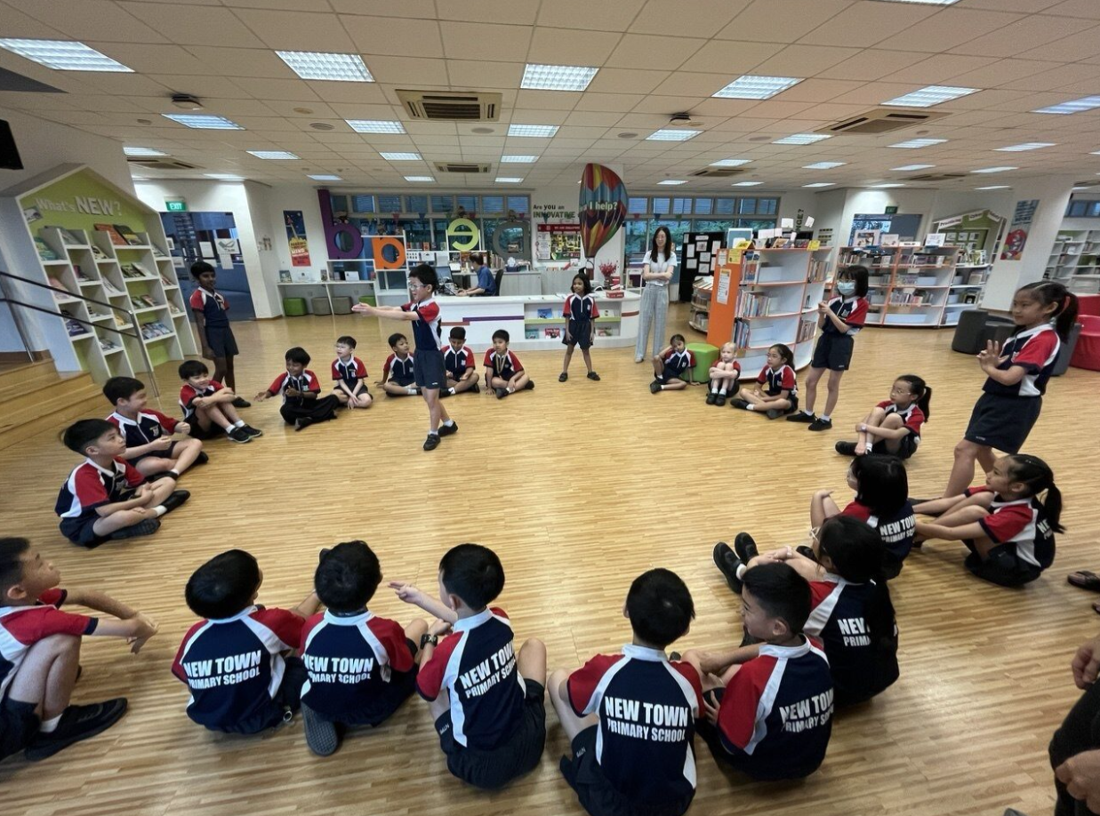 New Town Primary School students in uniform sit in a circle on a library floor while a student stands presenting.