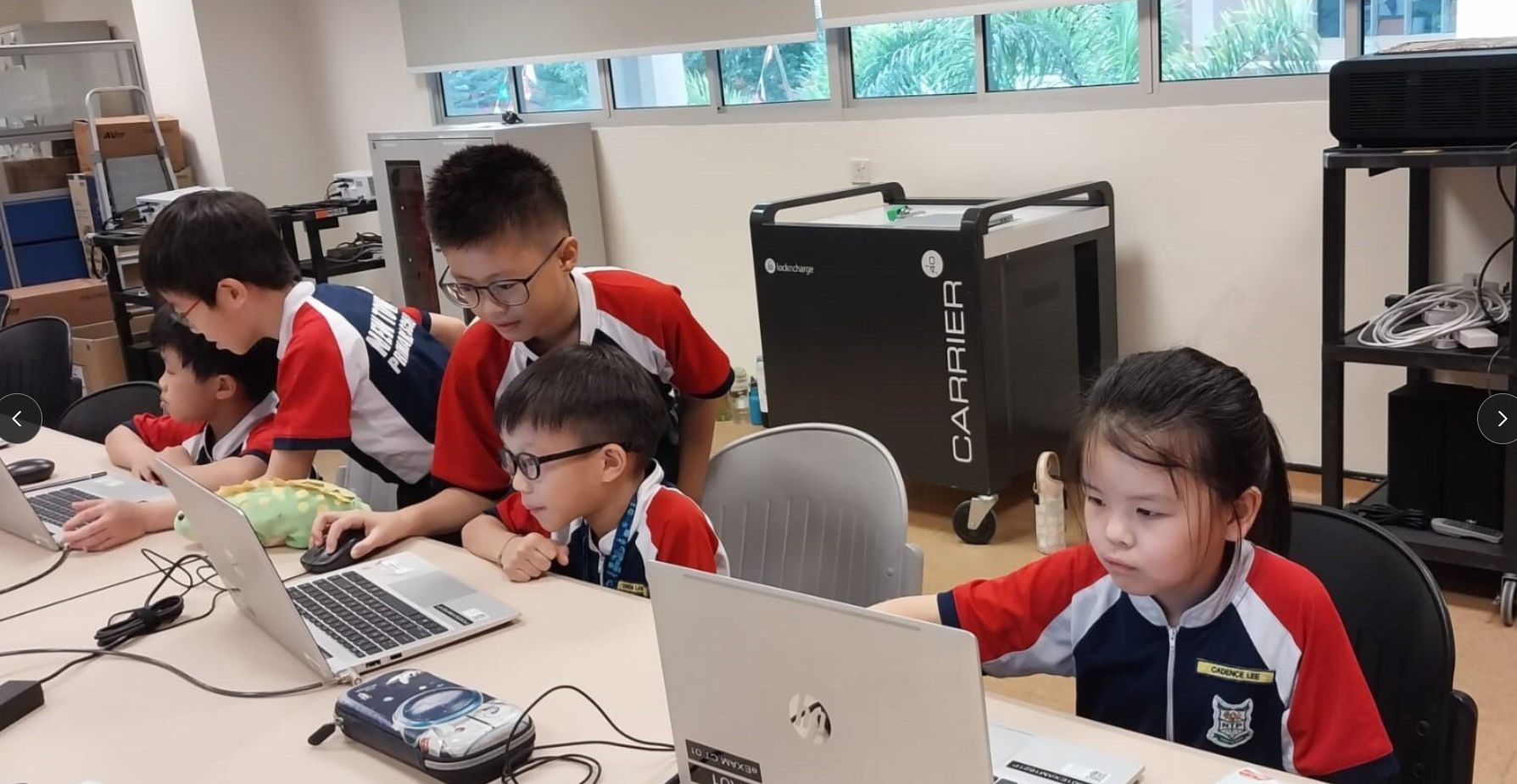 Four young students in red uniforms work on laptops at a table in a computer lab classroom.