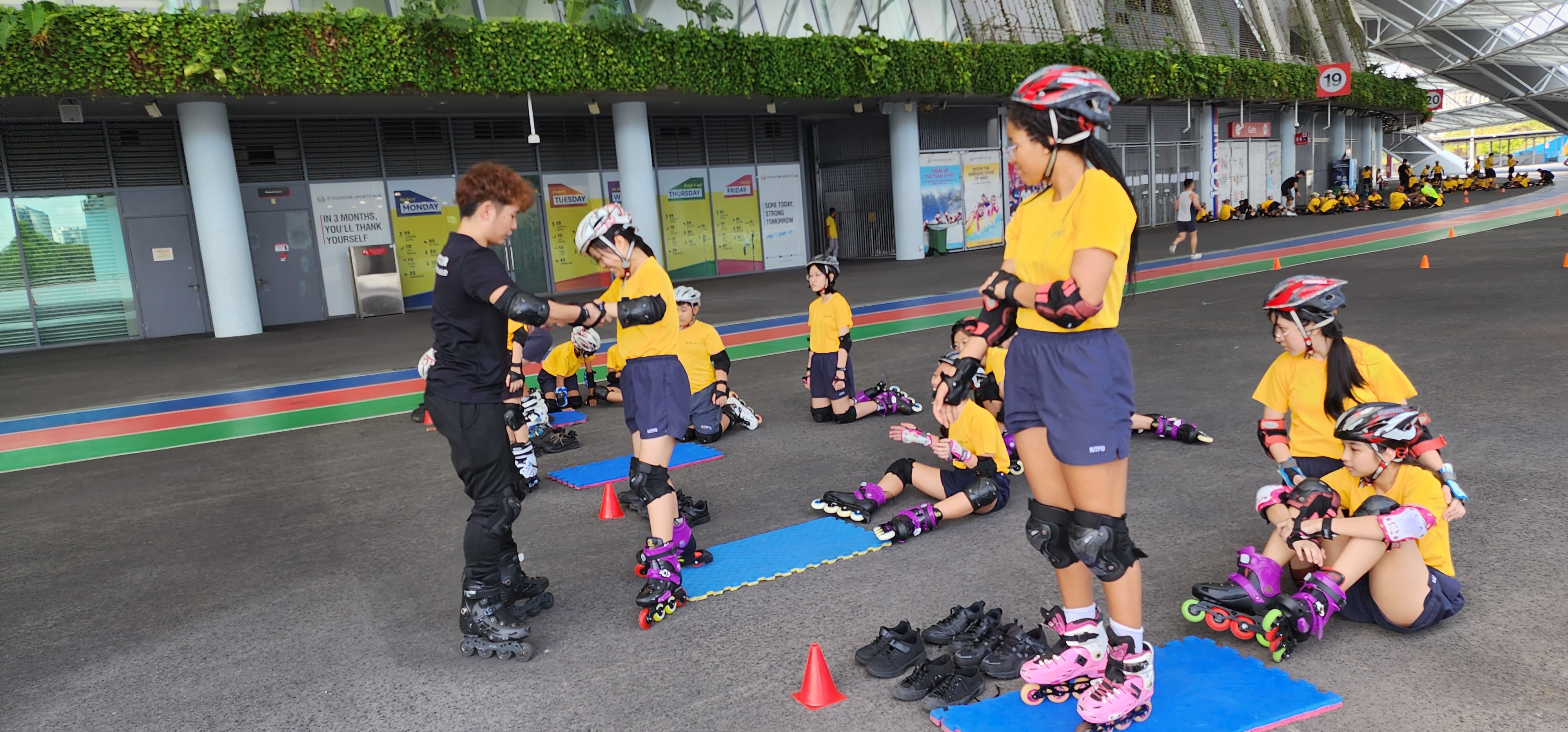 Children in yellow shirts and helmets learn inline skating on a track, guided by an instructor in black.