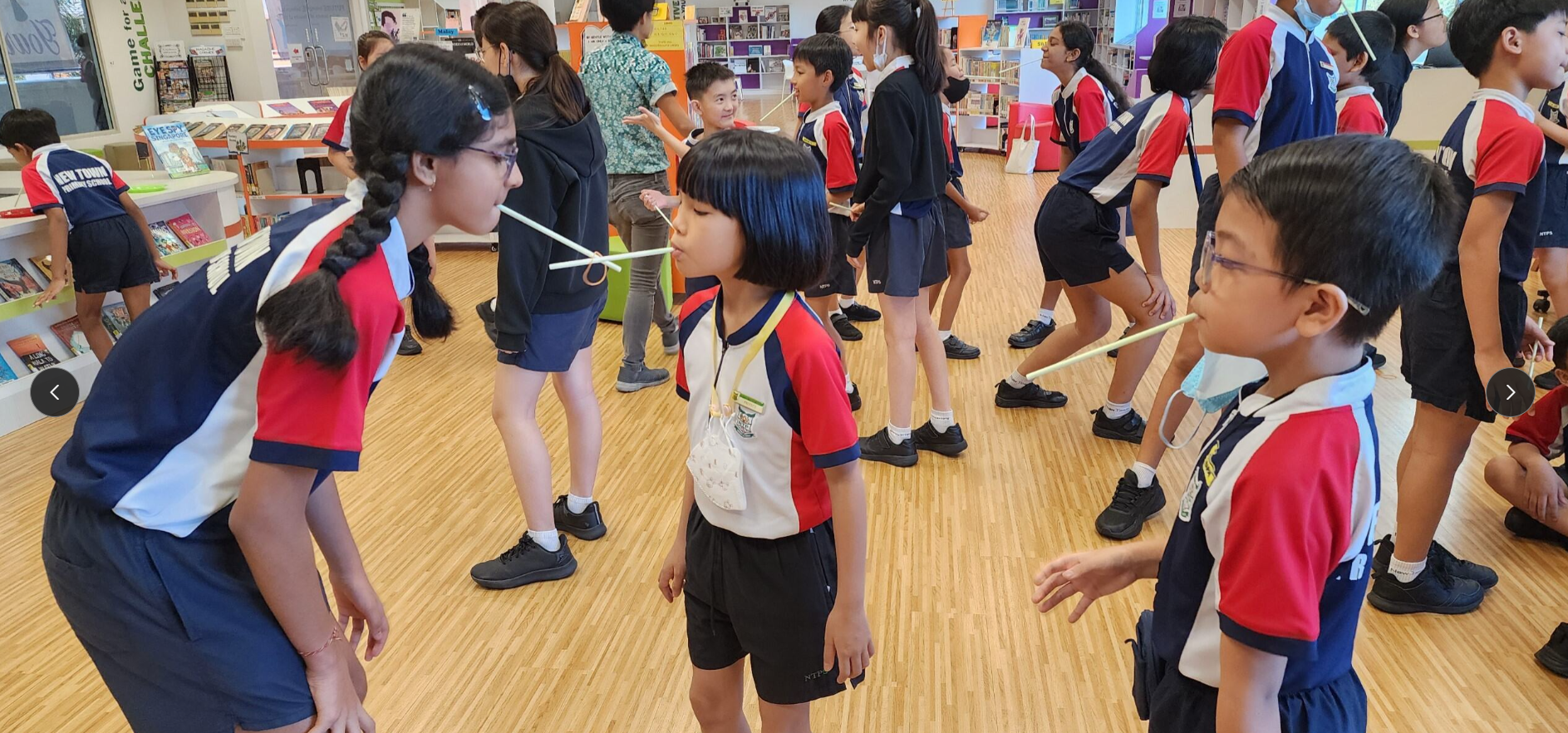 School children in uniforms balance straws between their mouths in a library activity.
