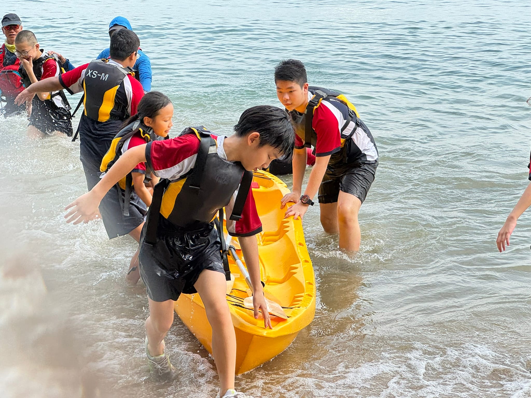 Several youths in life jackets carry a yellow kayak through shallow ocean waves near a shore.