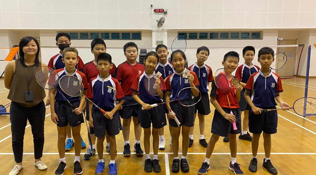 A group of young students in school uniforms hold badminton rackets indoors on a court, posing with an adult woman.