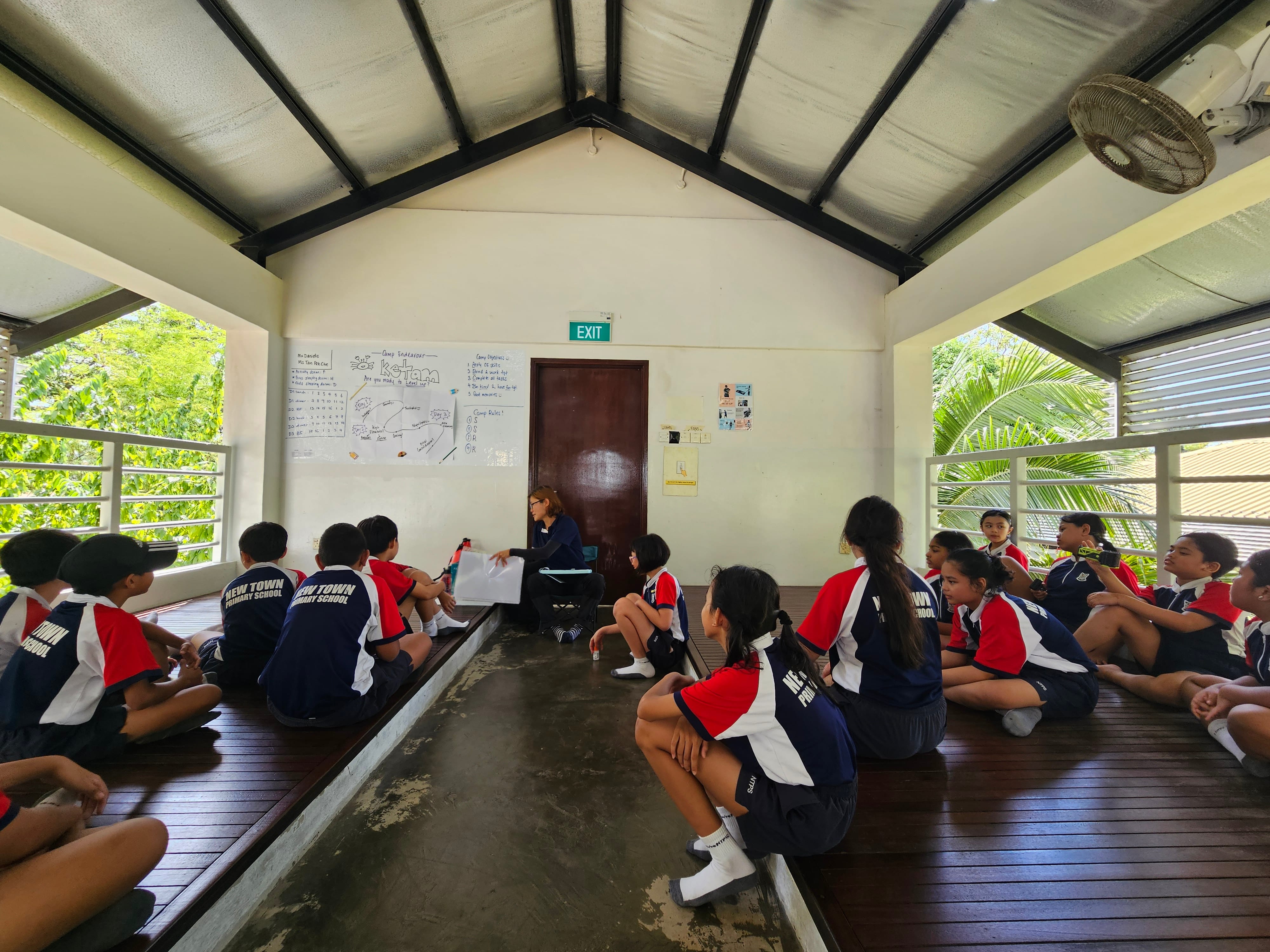 Primary school students in uniforms sit on a wooden floor in an open pavilion, facing a teacher with a whiteboard.